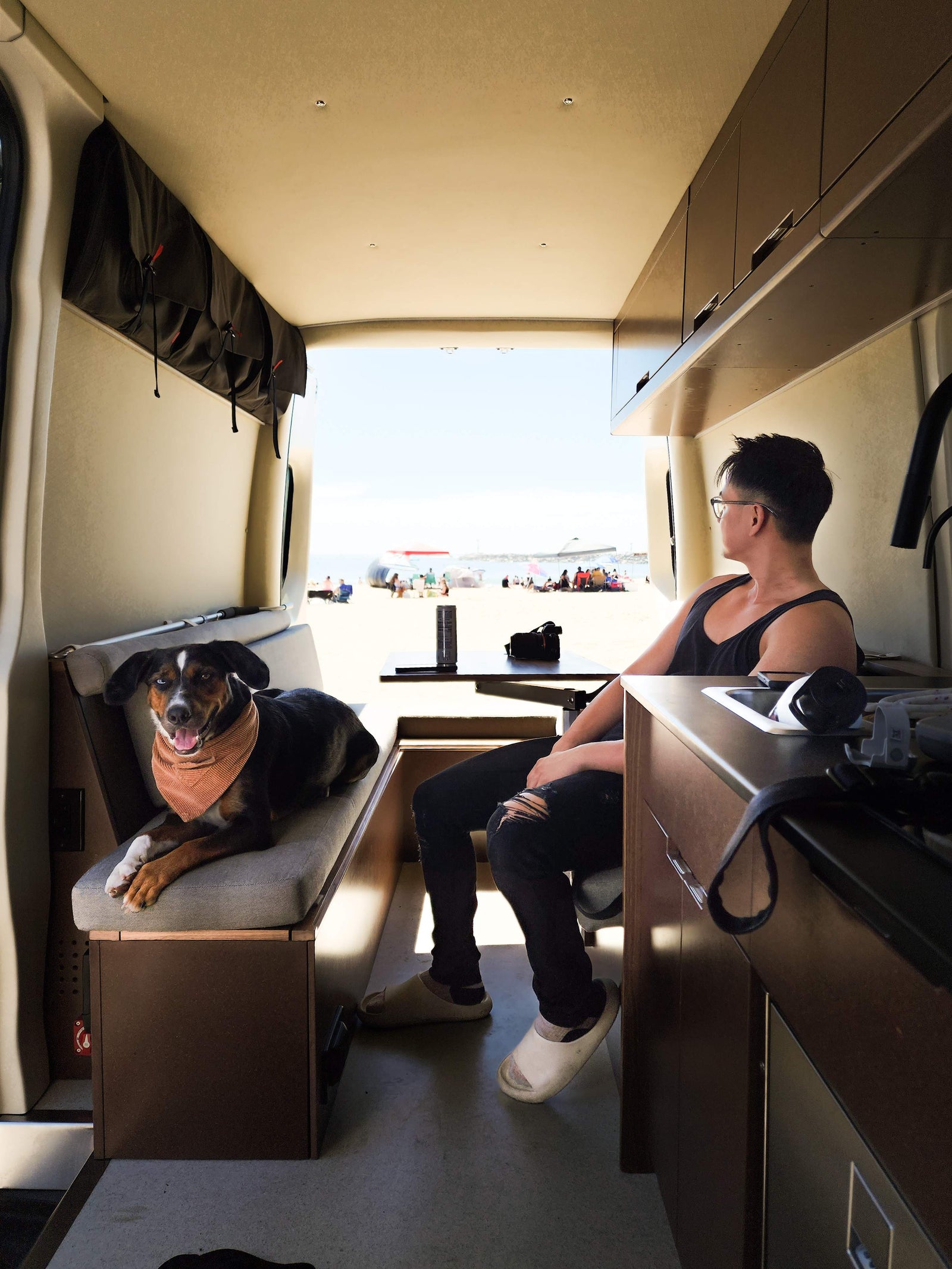 Inside an RV rental: A man and dog enjoy beach views. Overhead storage & kitchen are visible, with people on the sand outside.