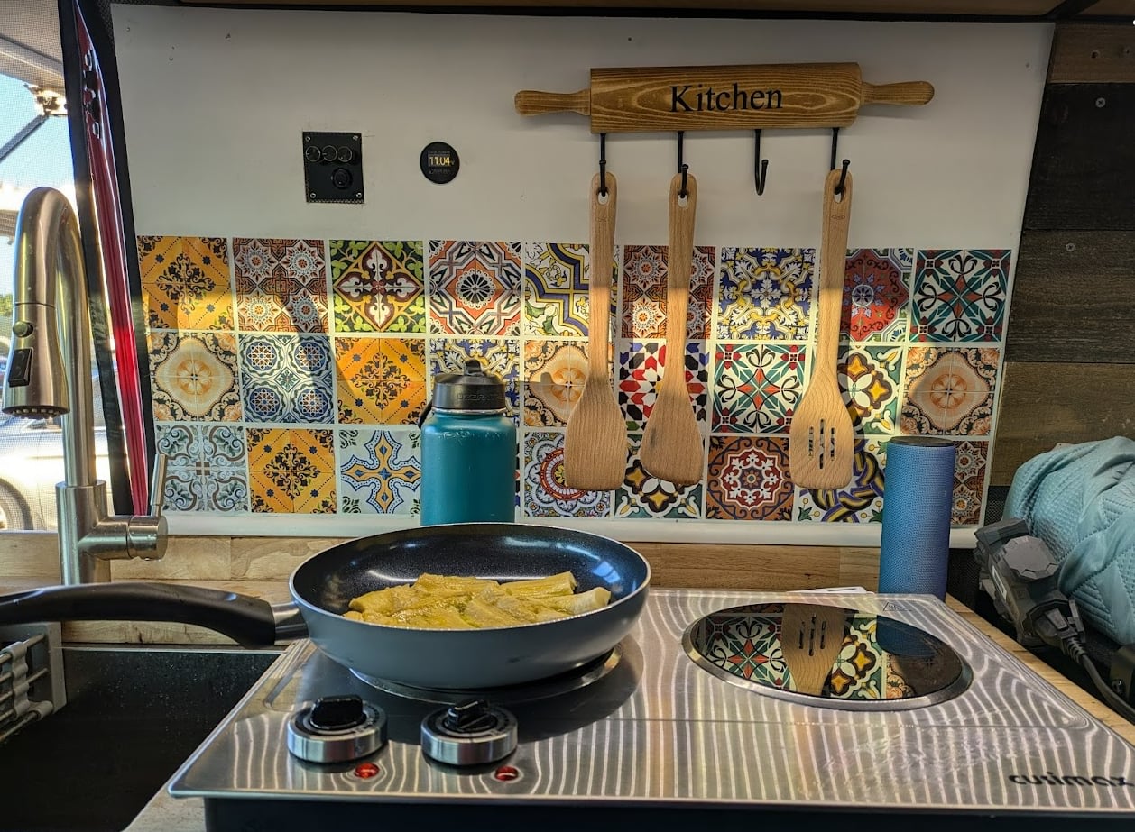 Inside an RV rental: a bright kitchen with a tiled backsplash, induction cooktop, wooden spatulas, and a pan of food.