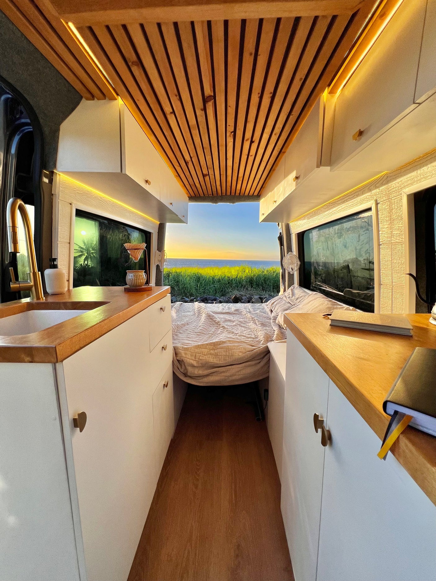 Cozy RV rental interior with wooden ceiling, functional kitchen & stunning ocean sunset view from the back. Perfect for van life adventures!