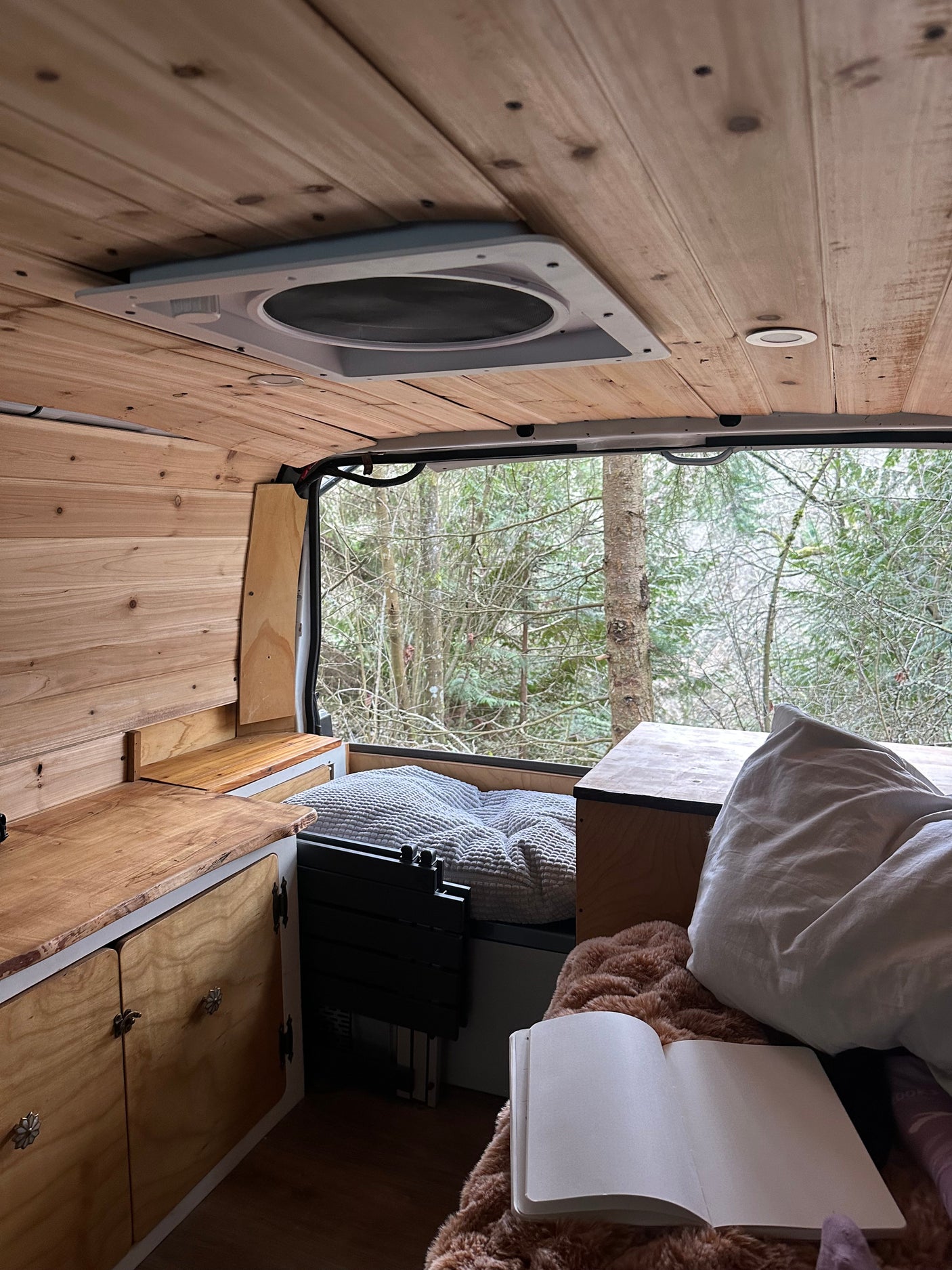 Cozy camper van RV rental interior with wooden walls & ceiling. Bed, kitchen area, & open notebook overlook a wintry forest view.