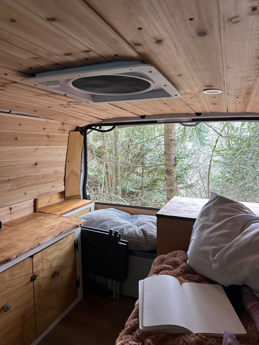 Cozy camper van RV rental interior with wooden walls & ceiling. Bed, kitchen area, & open notebook overlook a wintry forest view.