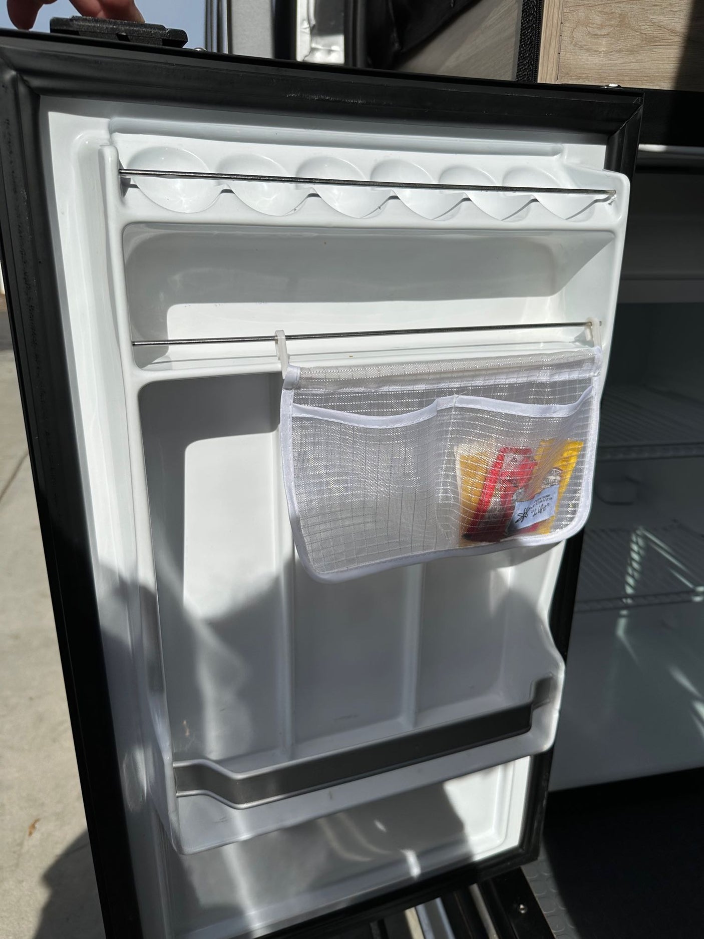Open RV rental refrigerator door showing internal shelving, an egg holder, and a mesh organizer pouch containing snacks.
