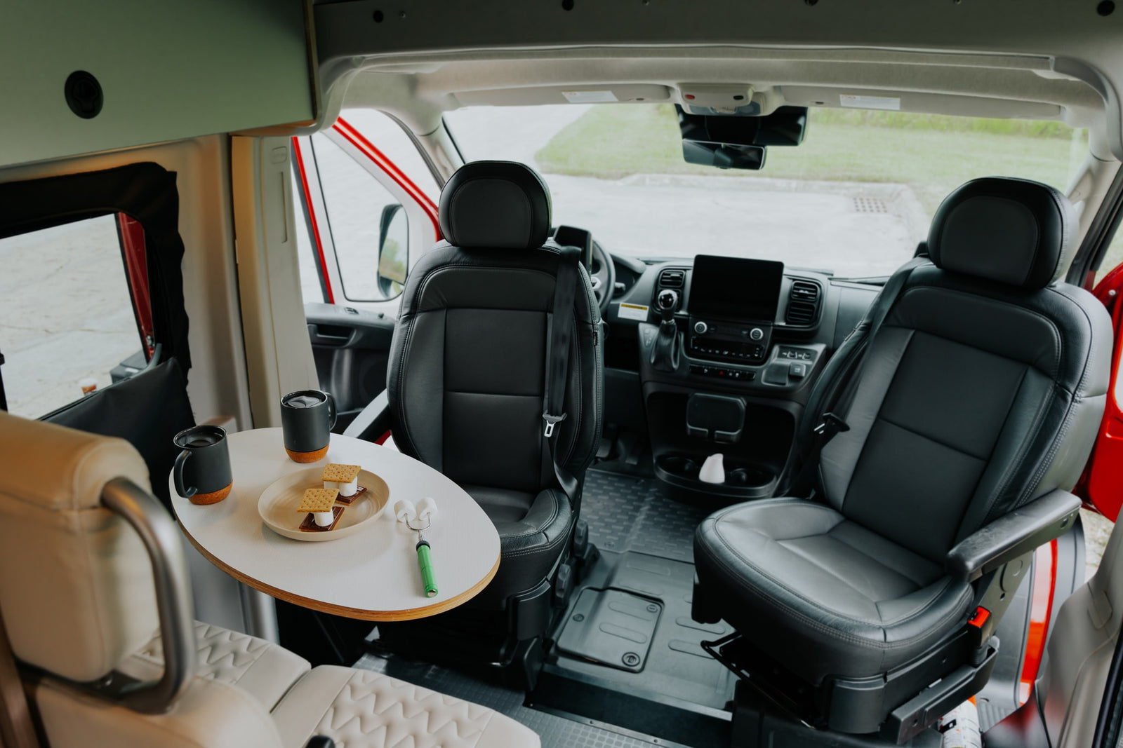 Comfortable RV rental interior looking toward the front: driver seats, dashboard, and table with s'mores & mugs.
