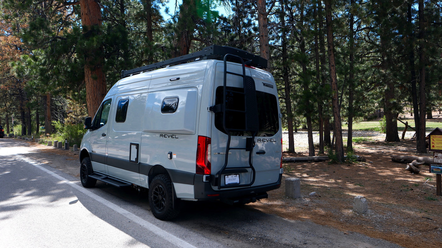 Grey Revel rv rental van parked in a sunny pine forest, featuring a roof rack and ladder, ready for outdoor adventures.