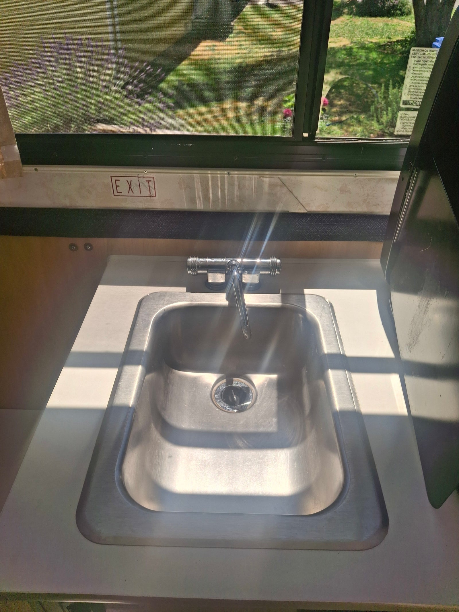 Bright RV rental interior: sink, faucet, and an "EXIT" sign. Outside, lavender plants visible through the window.