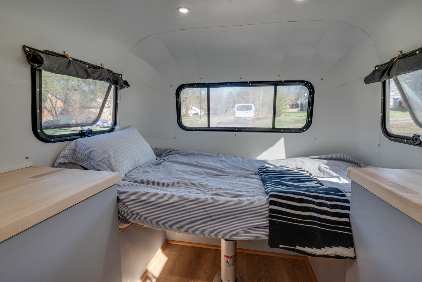 Interior view of a minimalist RV rental: a cozy bed with striped bedding, windows showing an outdoor scene, and light wood counters.