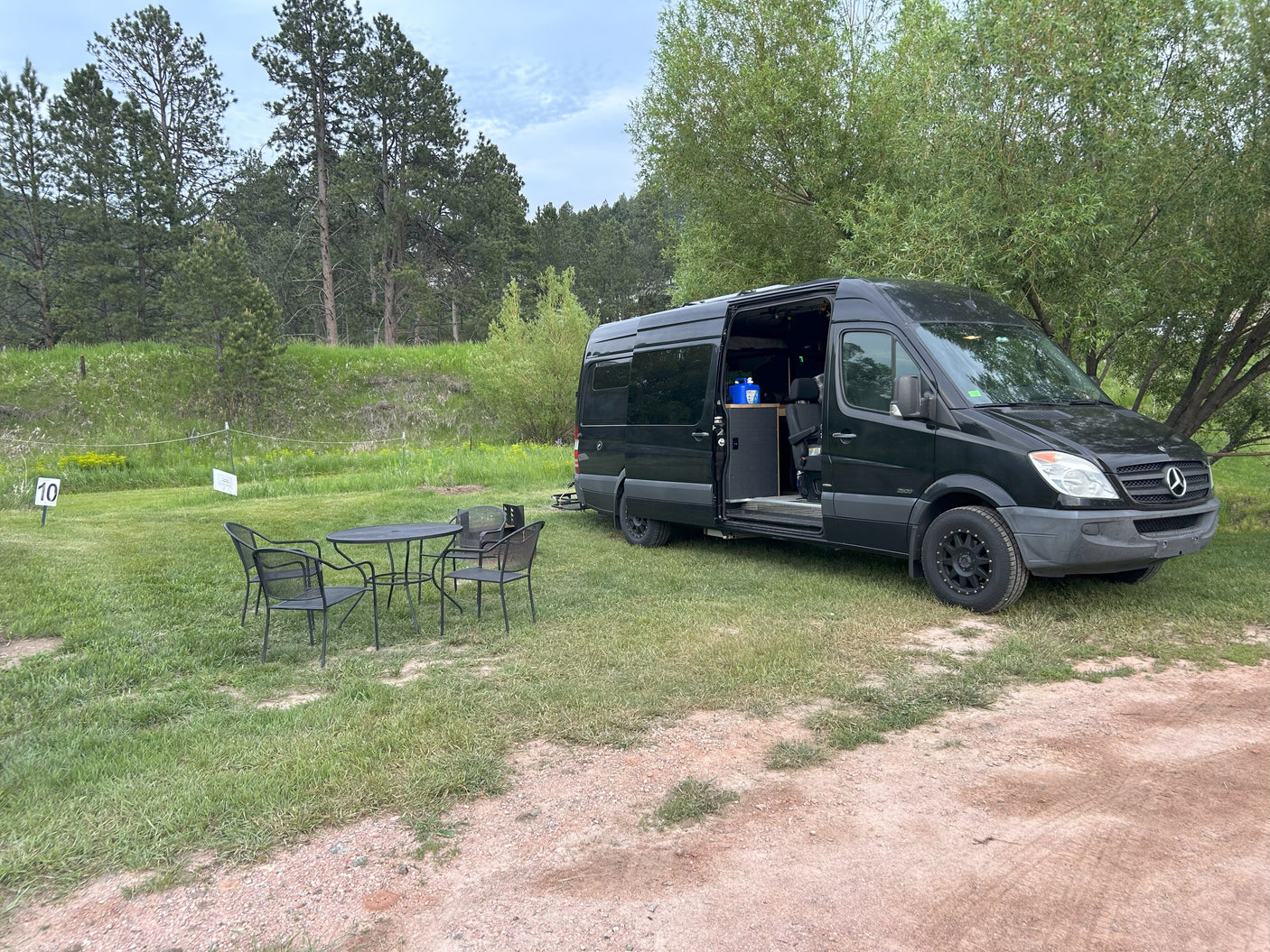 A black Sprinter van rv rental is parked at site 10 with outdoor seating and lush trees.