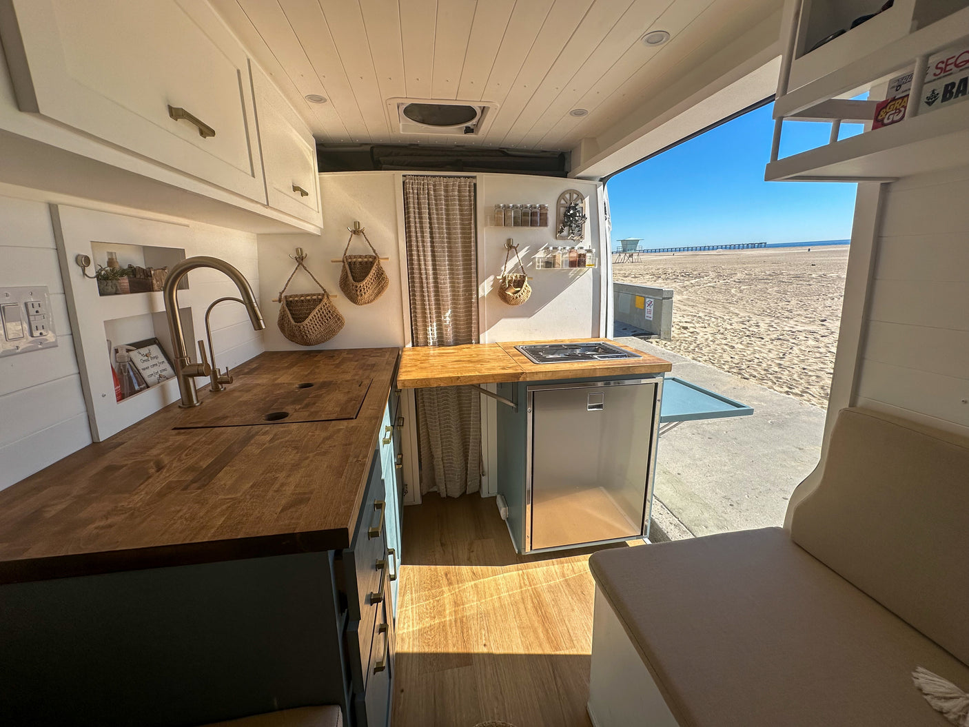 RV rental: Enjoy a bright camper van kitchen with wooden counters and fridge, open to a stunning beach and ocean view.