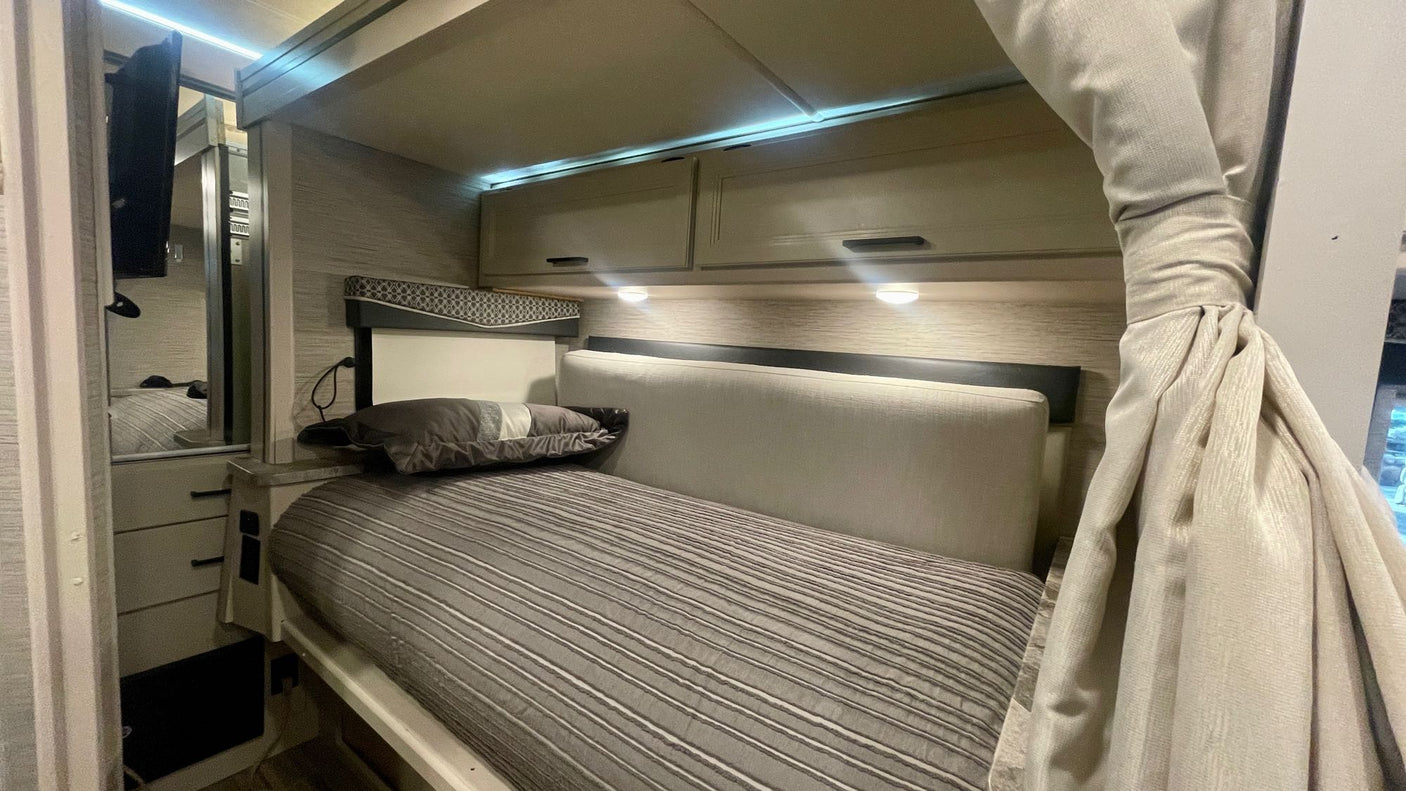 Cozy RV rental bedroom with comfortable bed, overhead storage, and integrated TV/mirror setup.