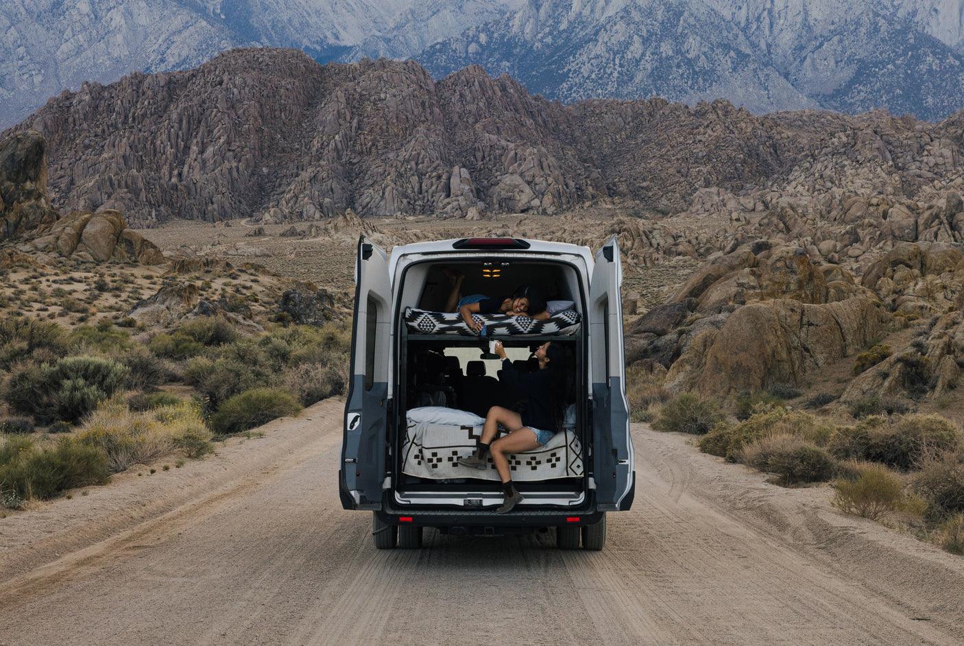 Relaxing in an **RV rental** camper van on a desert road. Two women enjoy their cozy bunks with rocky mountains beyond.