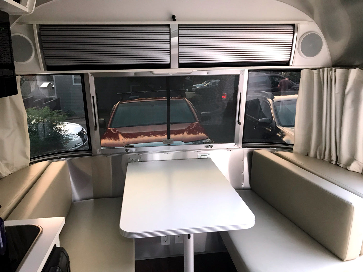 RV rental interior: A modern travel trailer dining area with a white table, light cushions, and large windows revealing parked vehicles outside.