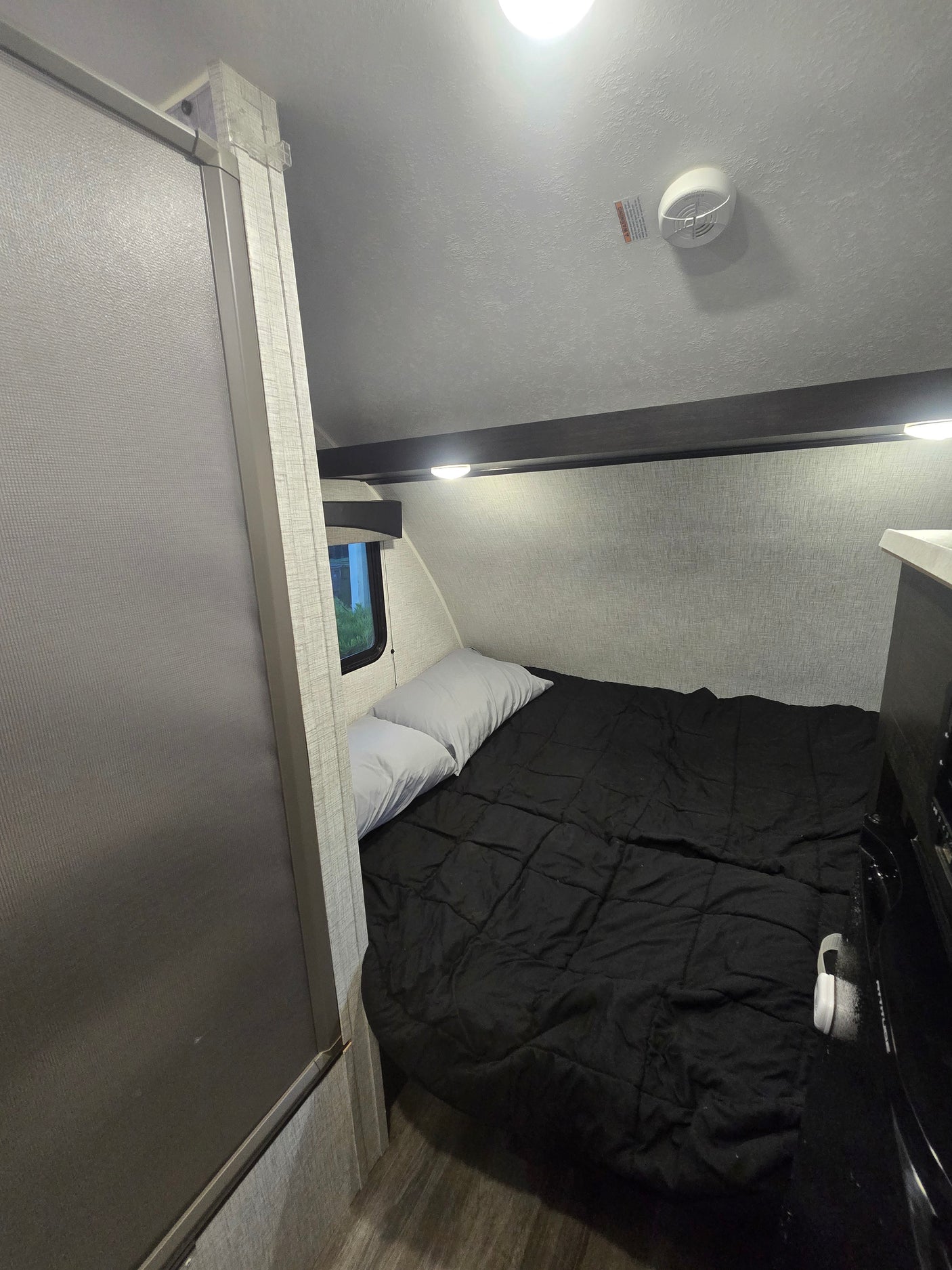 Compact RV rental bedroom with a bed, black comforter, and white pillows next to a window. Ceiling has lights and smoke detector.