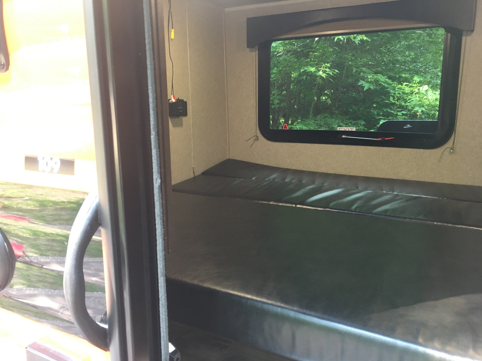 Inside an RV rental, a dark bunk bed faces a window with lush green trees. An exit sign and wall wiring are visible.