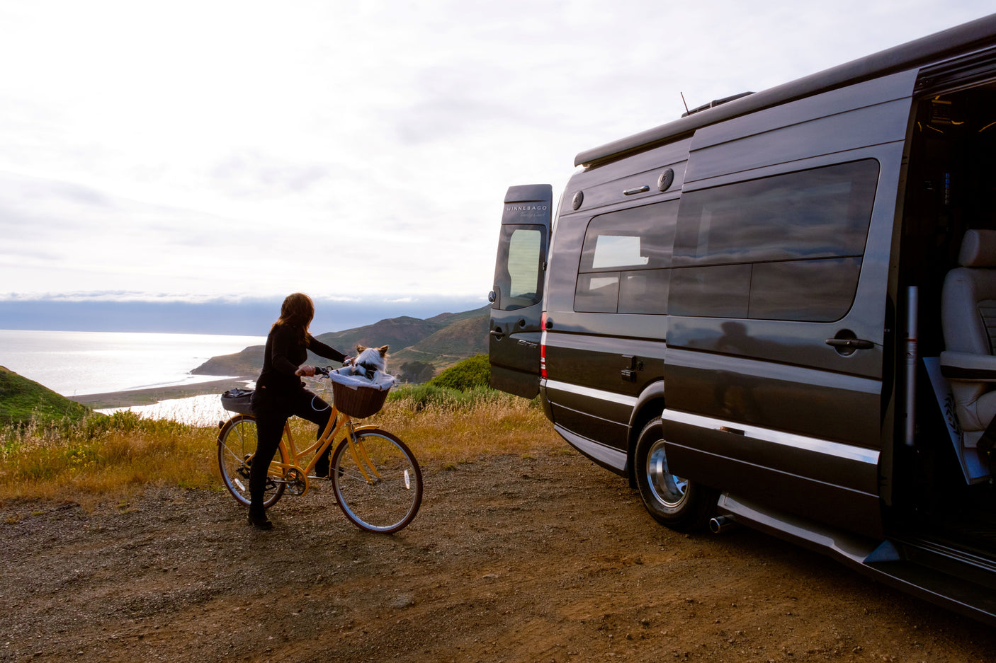 Scenic coastal RV rental journey: a dark gray Winnebago van parked as a person & dog enjoy a yellow bike ride.