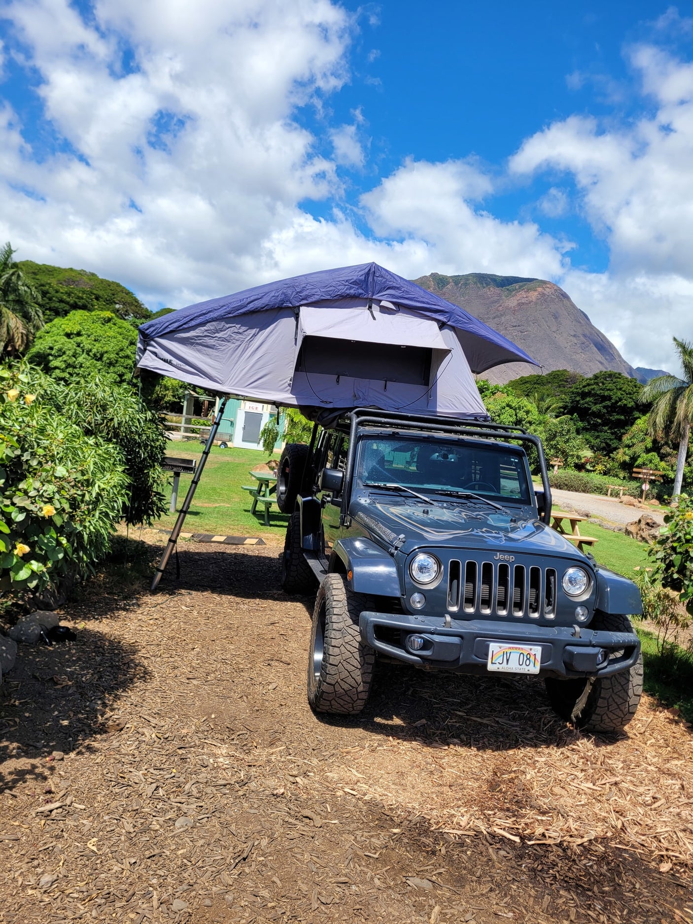 A dark gray Jeep Wrangler with a deployed rooftop tent offers a unique RV rental for camping amidst Hawaii's scenic beauty.