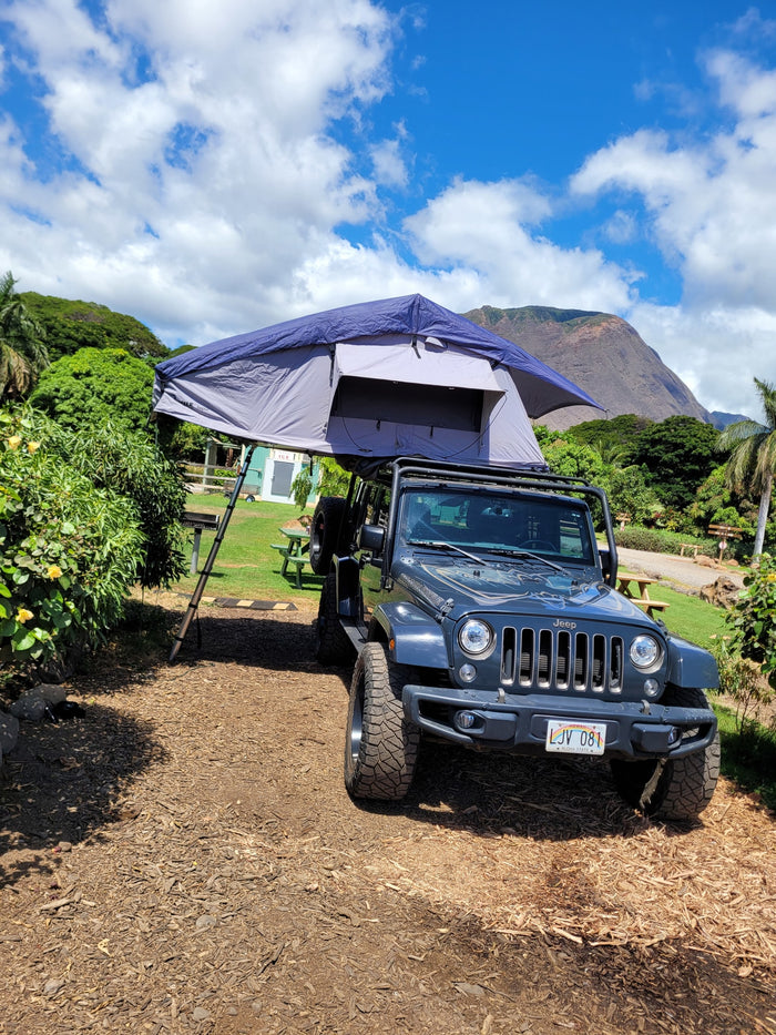 A dark gray Jeep Wrangler with a deployed rooftop tent offers a unique RV rental for camping amidst Hawaii's scenic beauty.