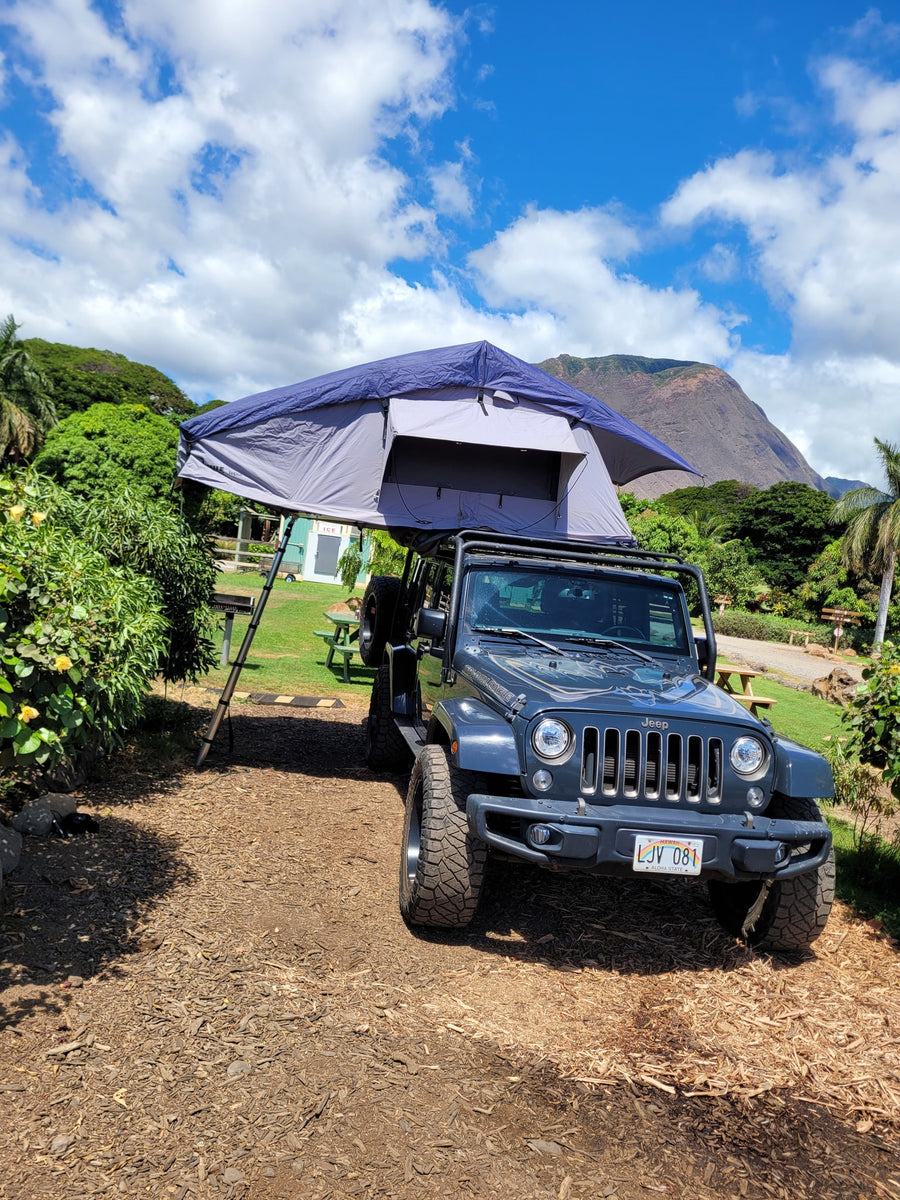 A dark gray Jeep Wrangler with a deployed rooftop tent offers a unique RV rental for camping amidst Hawaii's scenic beauty.