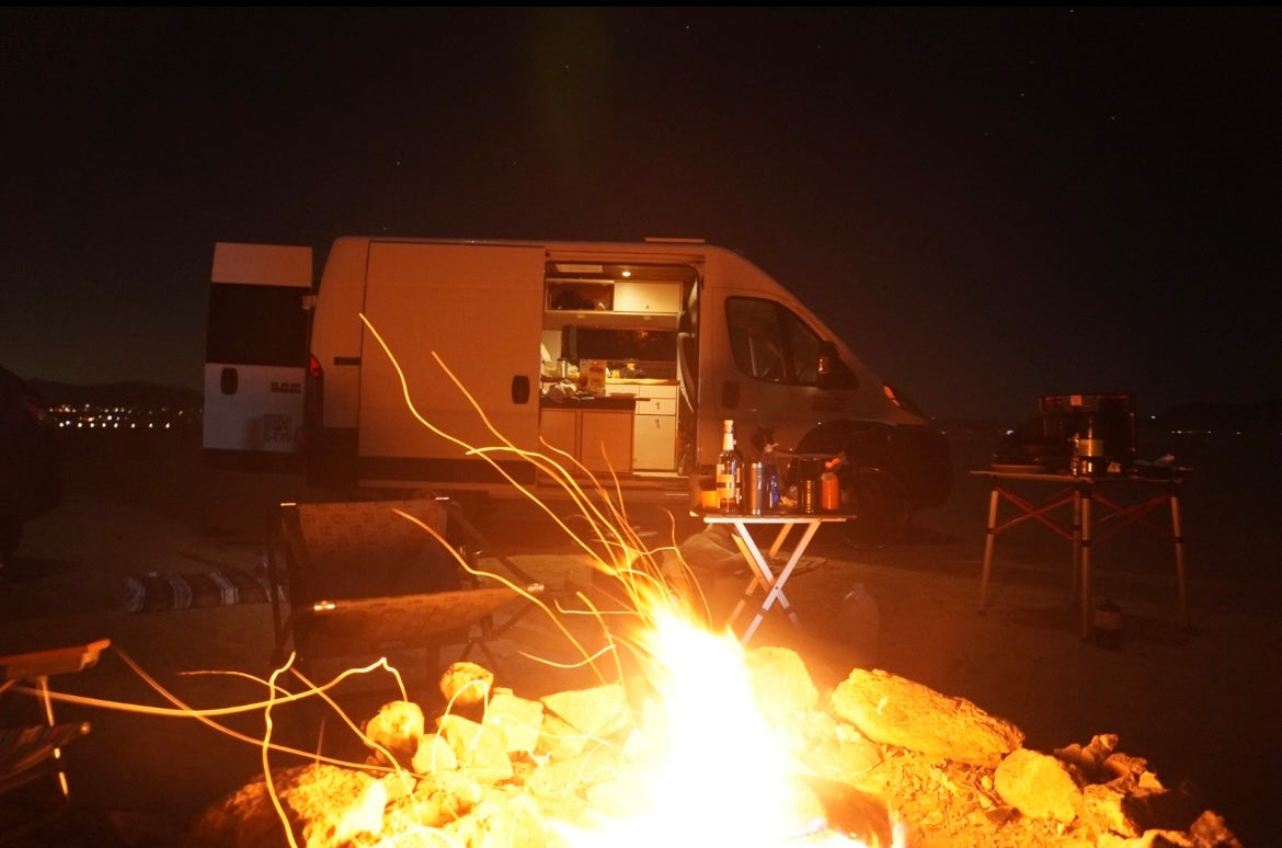 A cozy night under the stars: an RV rental camper van, vibrant campfire with sparks, and distant lights creating a serene outdoor escape.