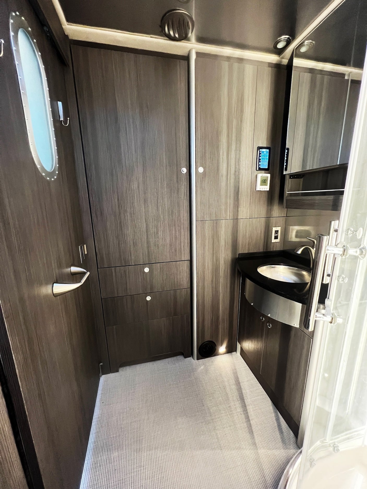 Modern RV rental washroom: dark wood-grain cabinets, sleek sink, mirror, shower, and a door with a porthole window.
