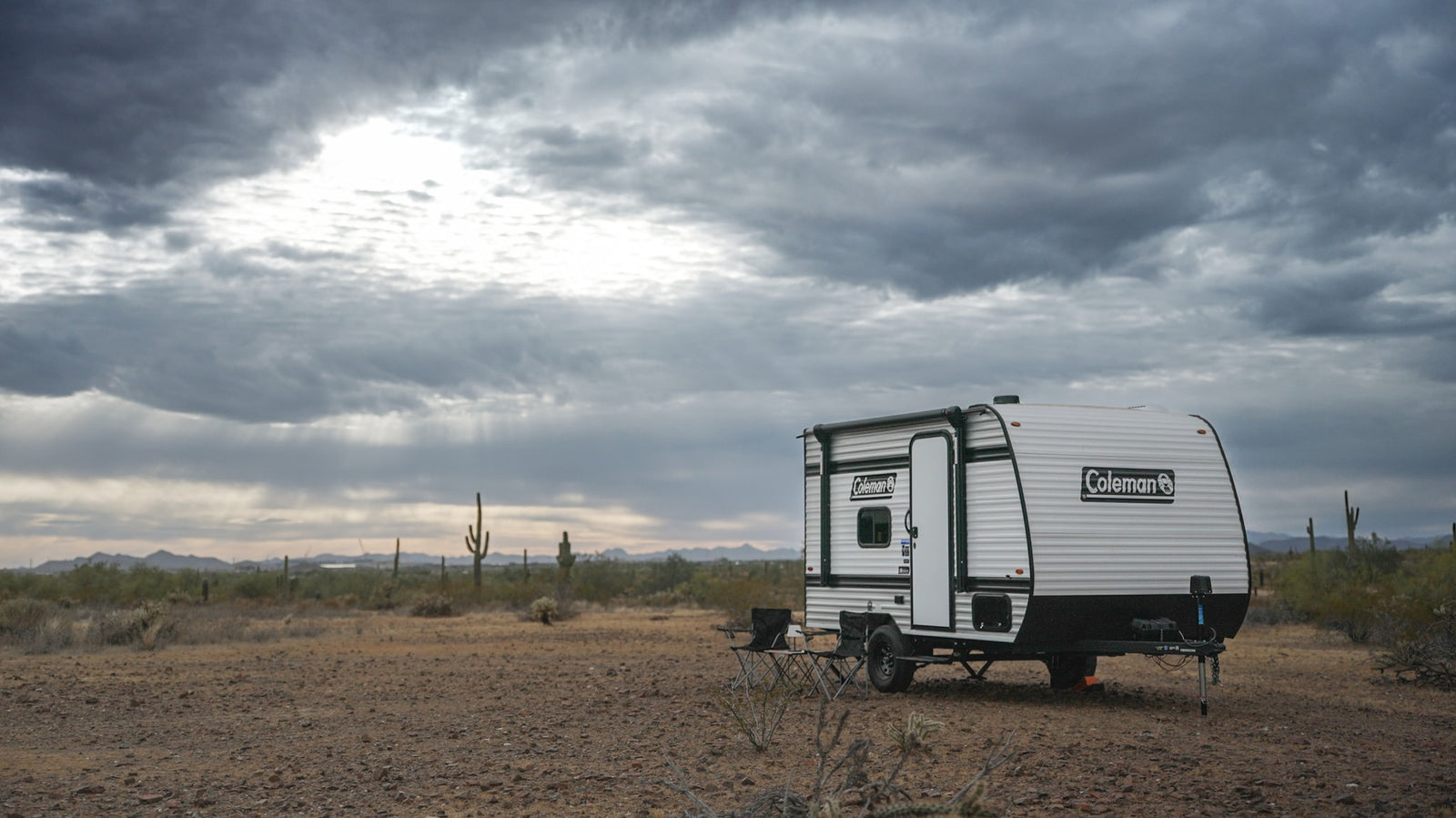 Coleman rv rental in a vast, rocky desert. A travel trailer with chairs set up, under a dramatic, cloudy sky with sun rays.