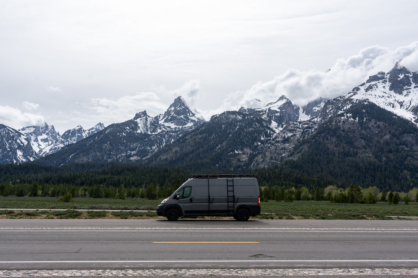 Stunning landscape with a grey RV rental van on a road, backed by impressive snow-capped Grand Teton mountains and forest.