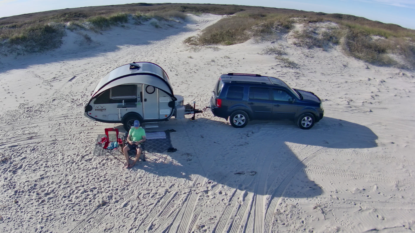 RV rental on a sandy beach. A person relaxes beside a teardrop trailer towed by an SUV, surrounded by dunes.