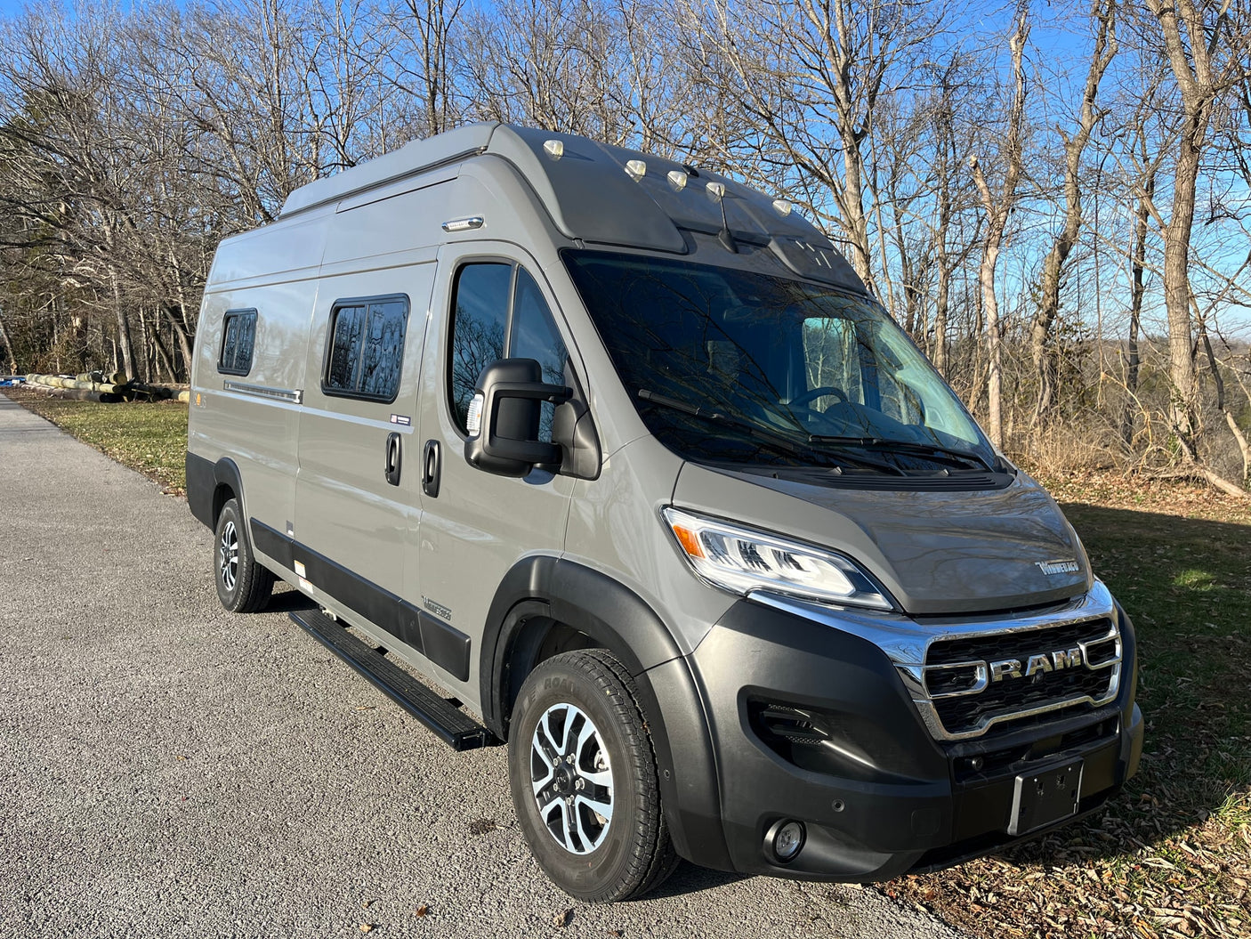 A modern gray Winnebago Solis camper van, a prime RV rental choice, parked outdoors on a bright, clear day among bare trees.