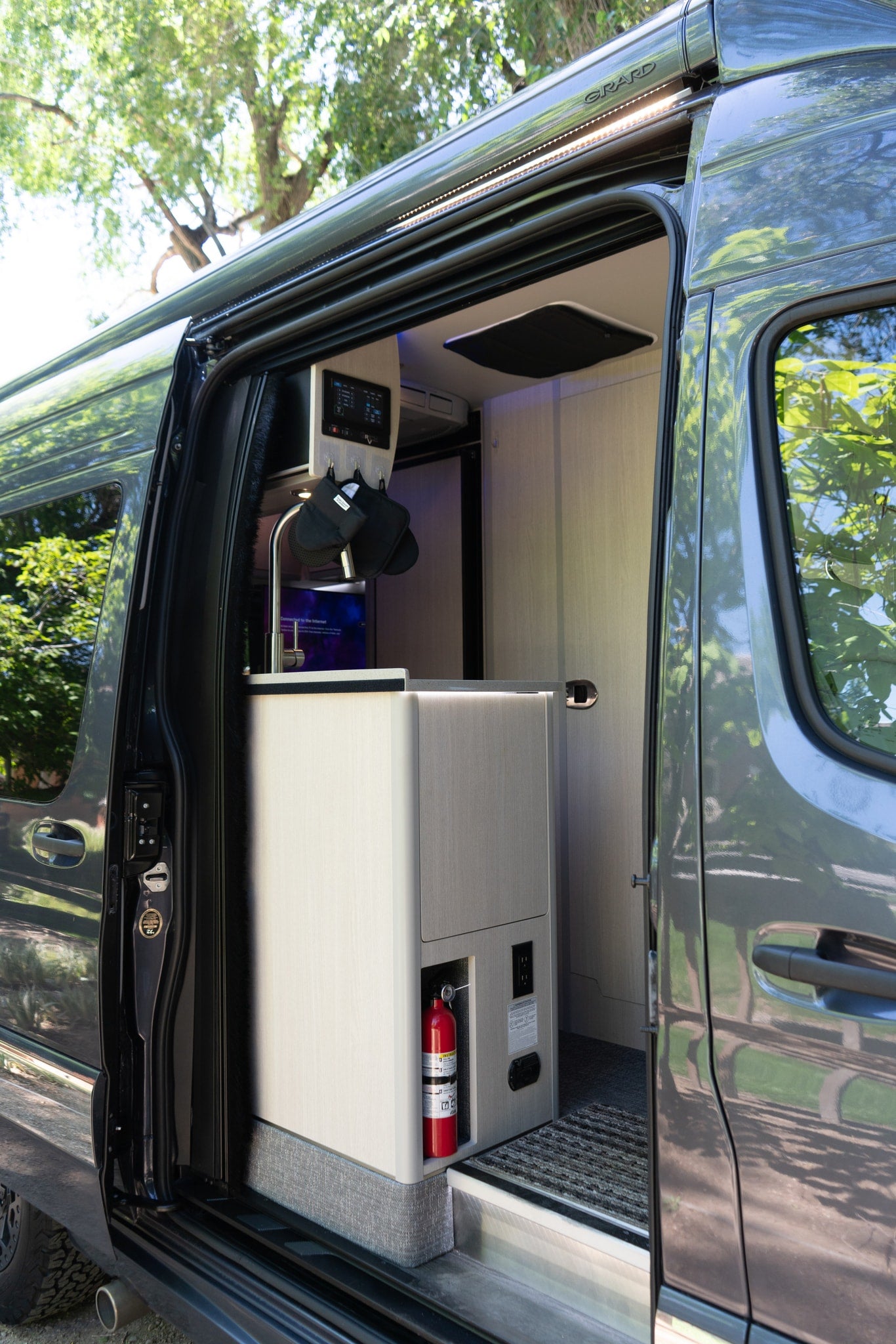 Modern RV rental interior: kitchen with sink, control panel, fire extinguisher, and outlets visible through the open camper van door. — photo 9
