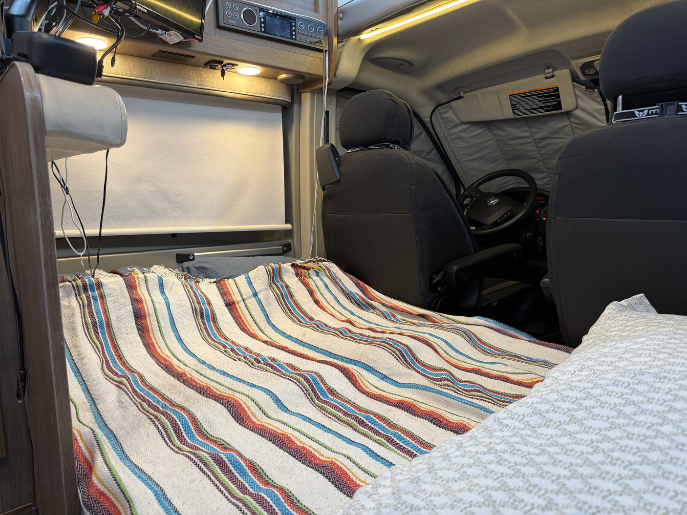 Cozy RV rental interior. Features a bed with a vibrant striped blanket, built-in entertainment, and front seats.