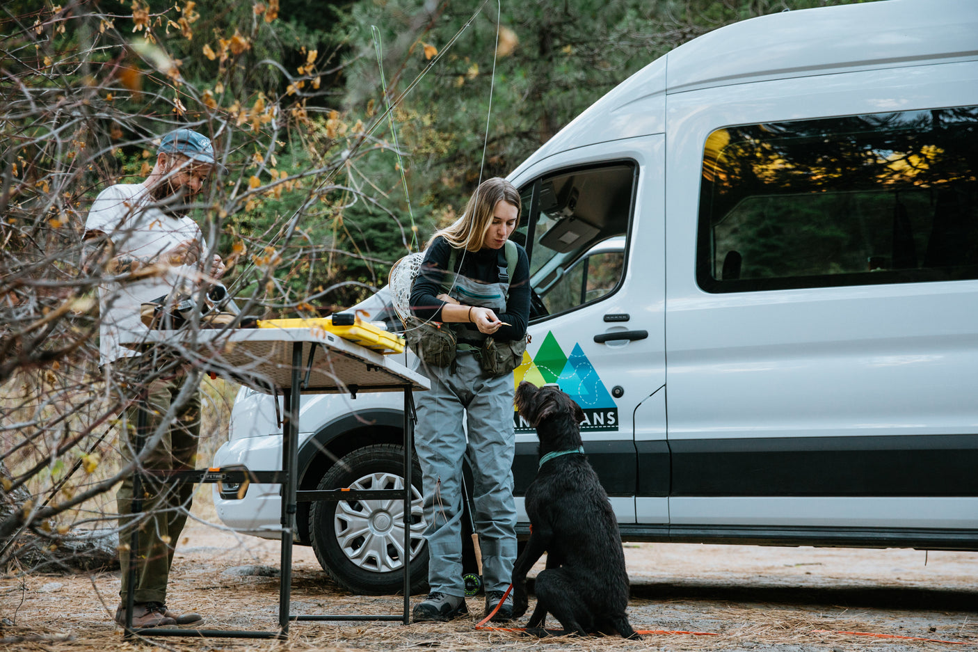 Two people and their dog set up for fly fishing from a white RV rental camper van in the woods.