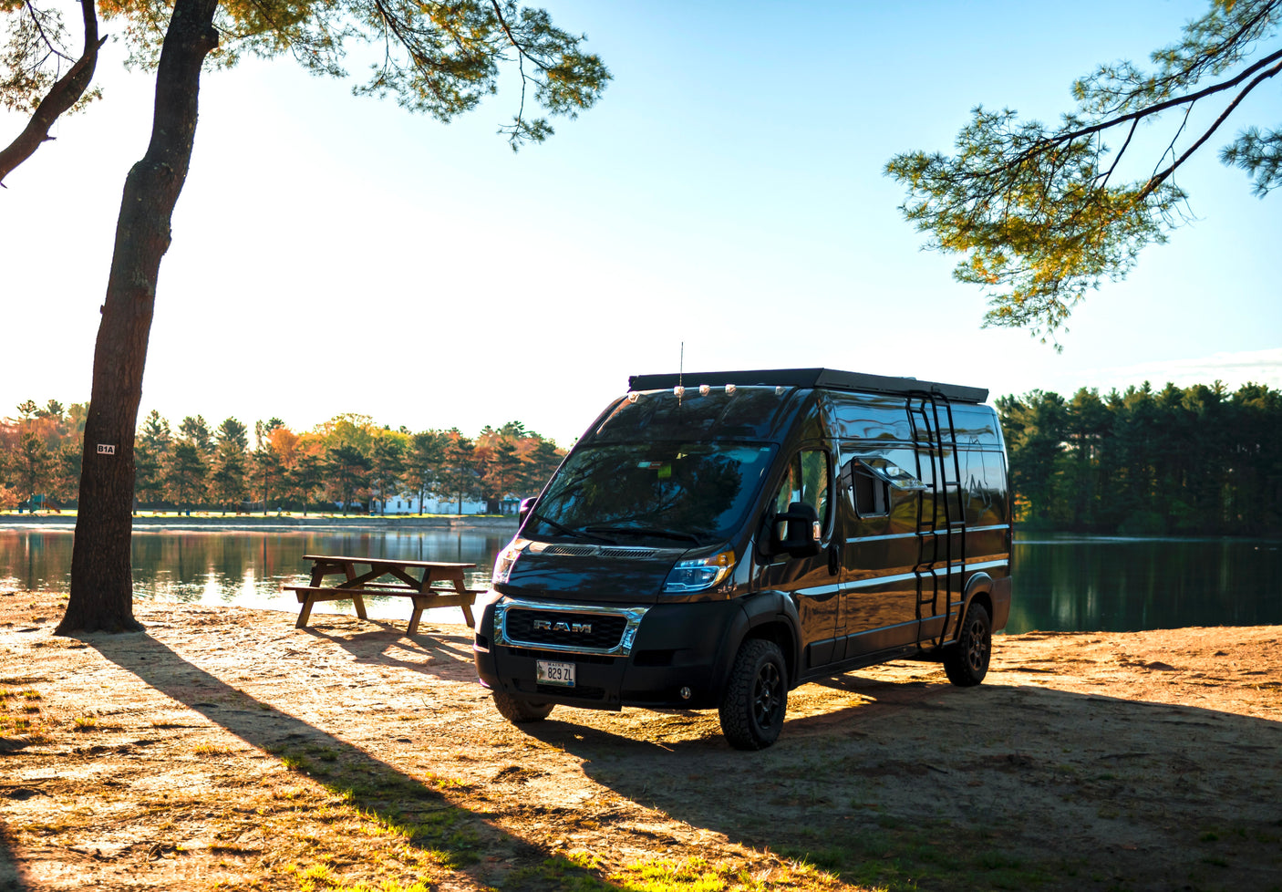 Black camper van RV rental parked lakeside, featuring a picnic table and trees. Enjoy serene nature and outdoor adventures under sunny skies.