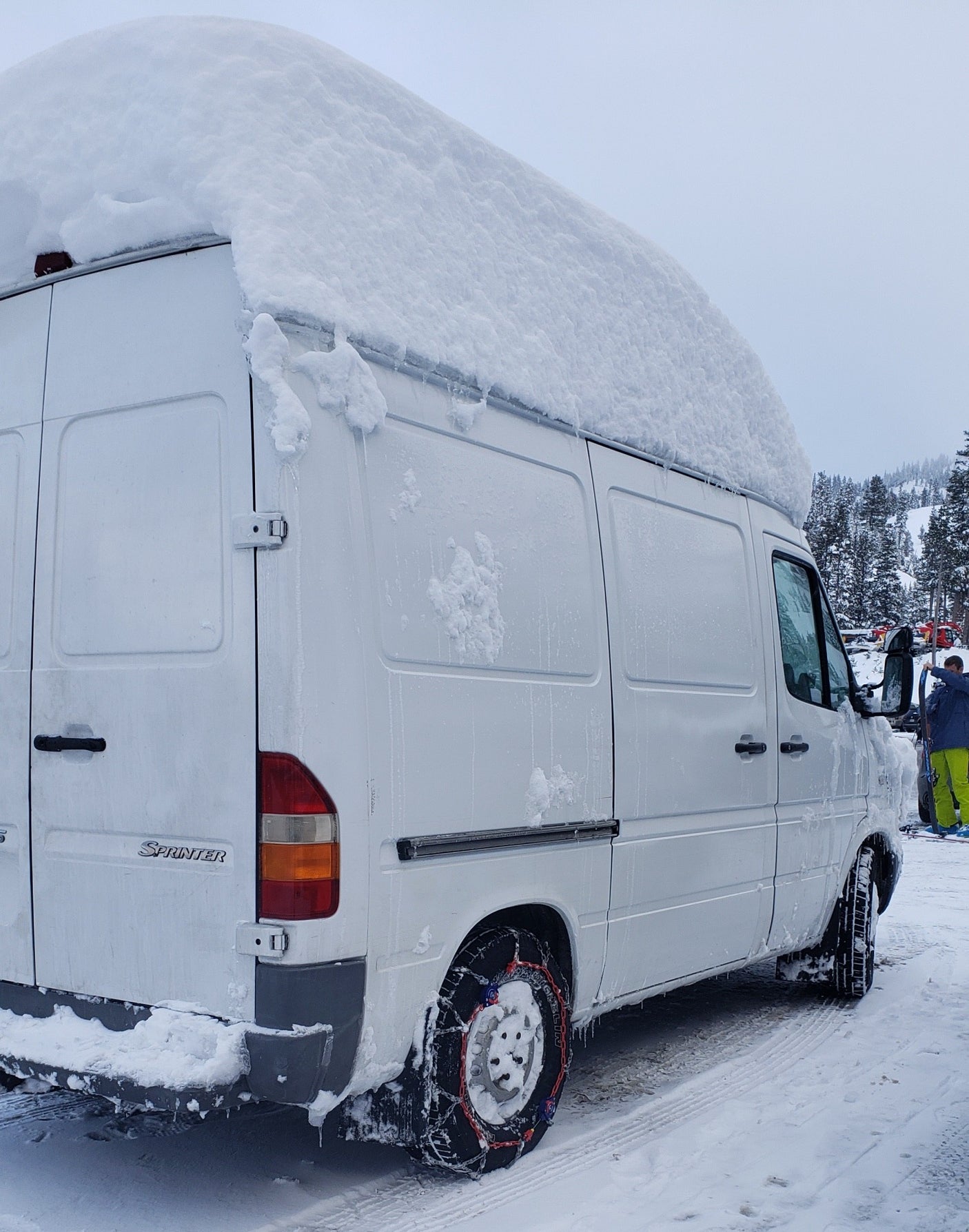 Heavy snow blankets this white Mercedes Sprinter van, ready for a winter RV rental adventure with tire chains.