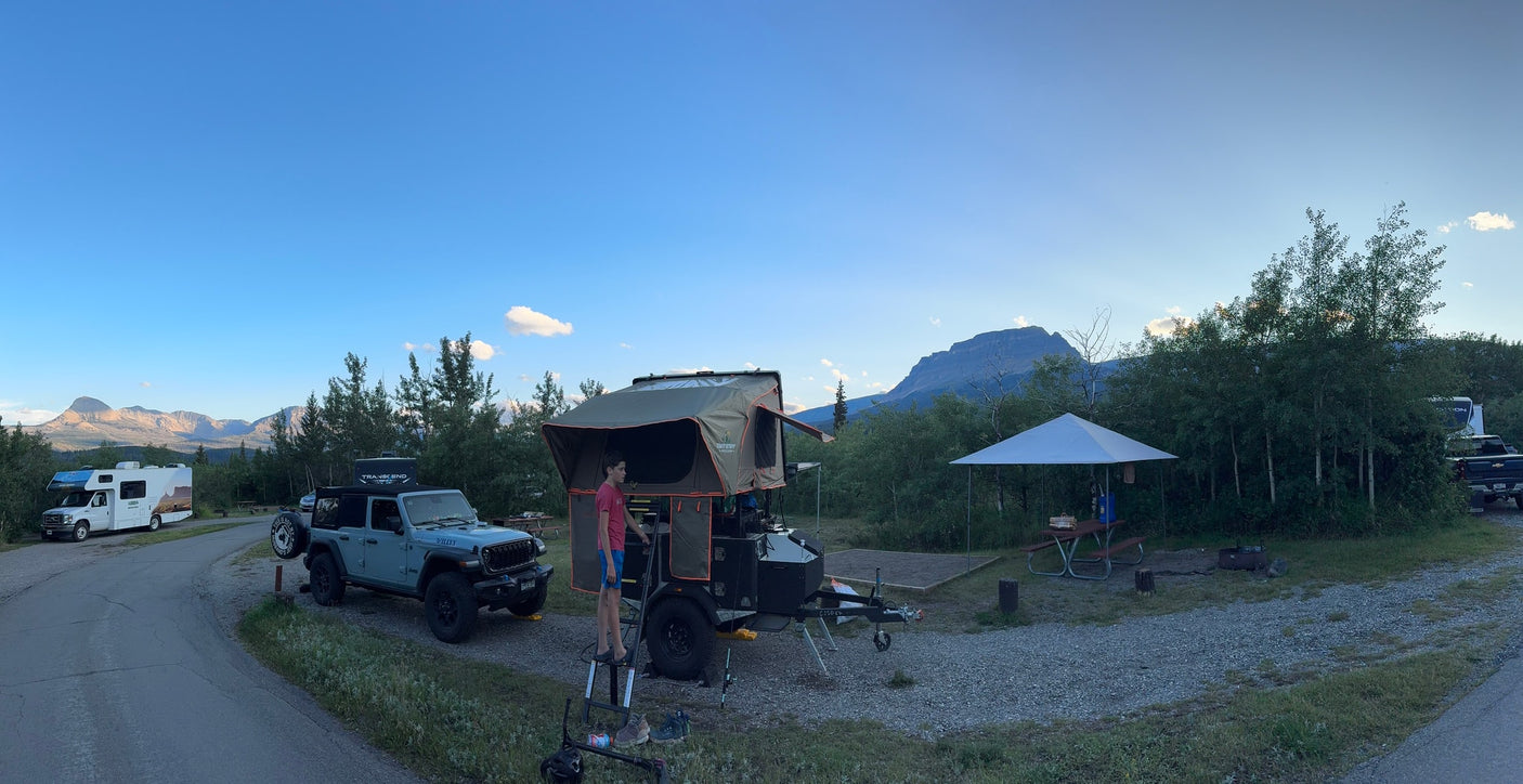 A campsite featuring a Jeep, a trailer with a rooftop tent, and an RV rental. Majestic mountains rise under a clear sky.