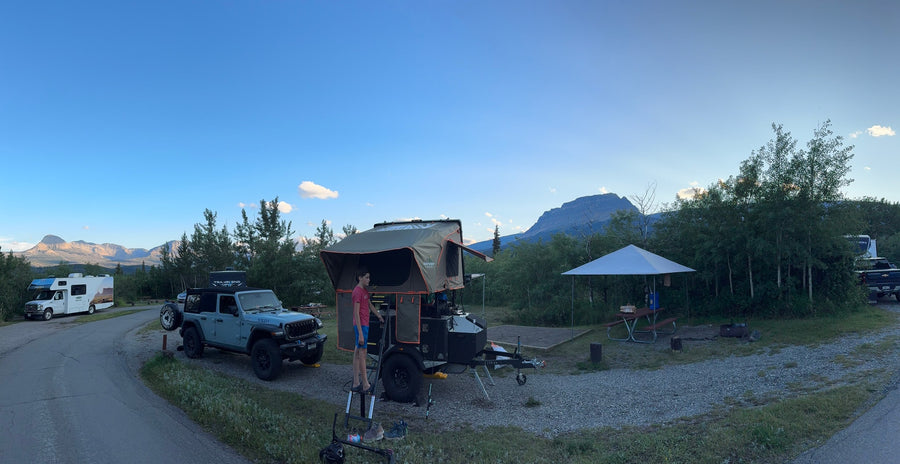 A campsite featuring a Jeep, a trailer with a rooftop tent, and an RV rental. Majestic mountains rise under a clear sky.