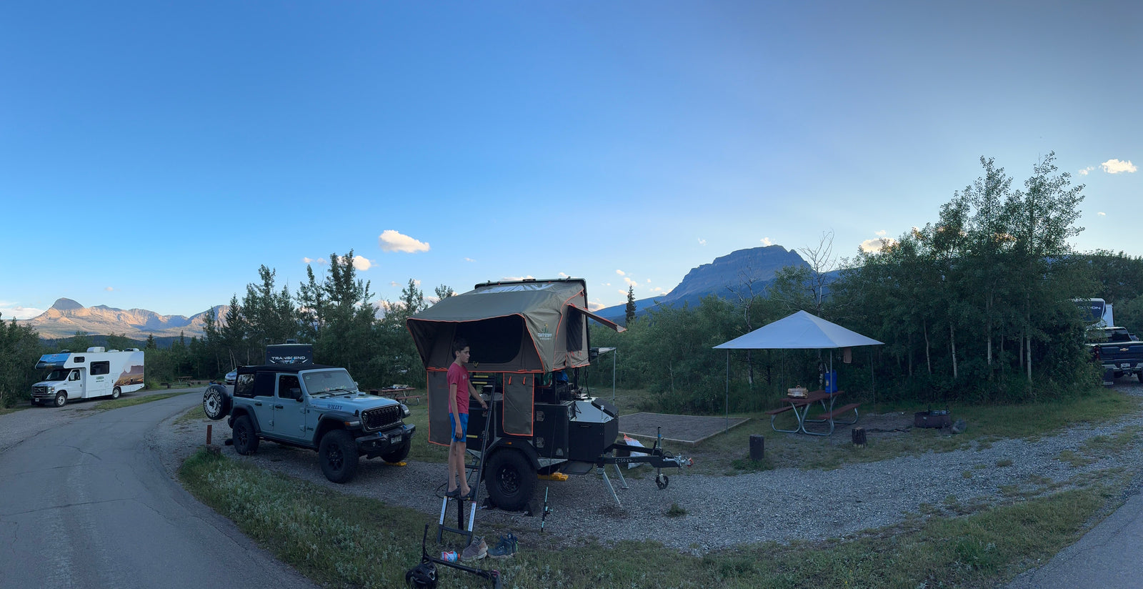 A campsite featuring a Jeep, a trailer with a rooftop tent, and an RV rental. Majestic mountains rise under a clear sky.