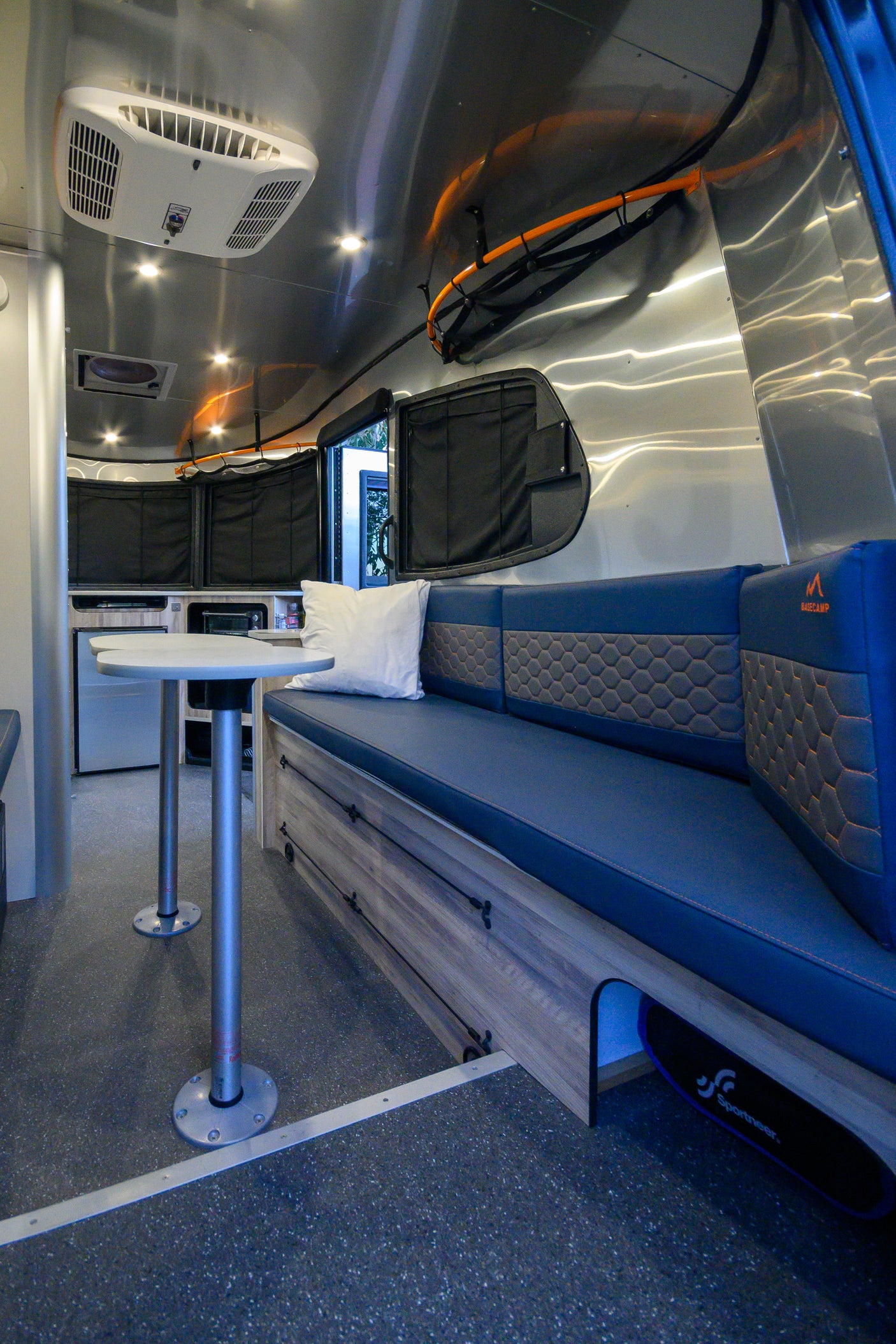 Modern rv rental interior: blue seating with storage, small table, polished aluminum walls, AC, and speckled floor.