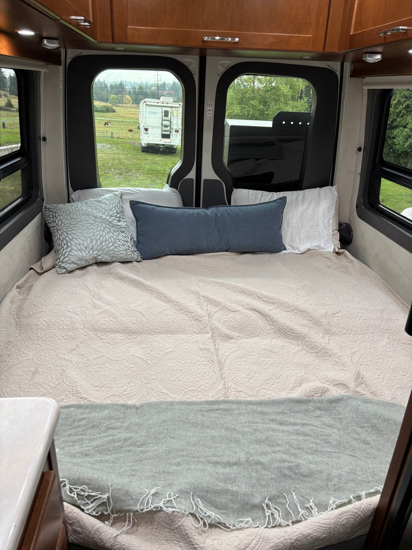 Cozy camper van bed for RV rental, featuring soft quilt, pillows, and serene views of a field and distant RV.