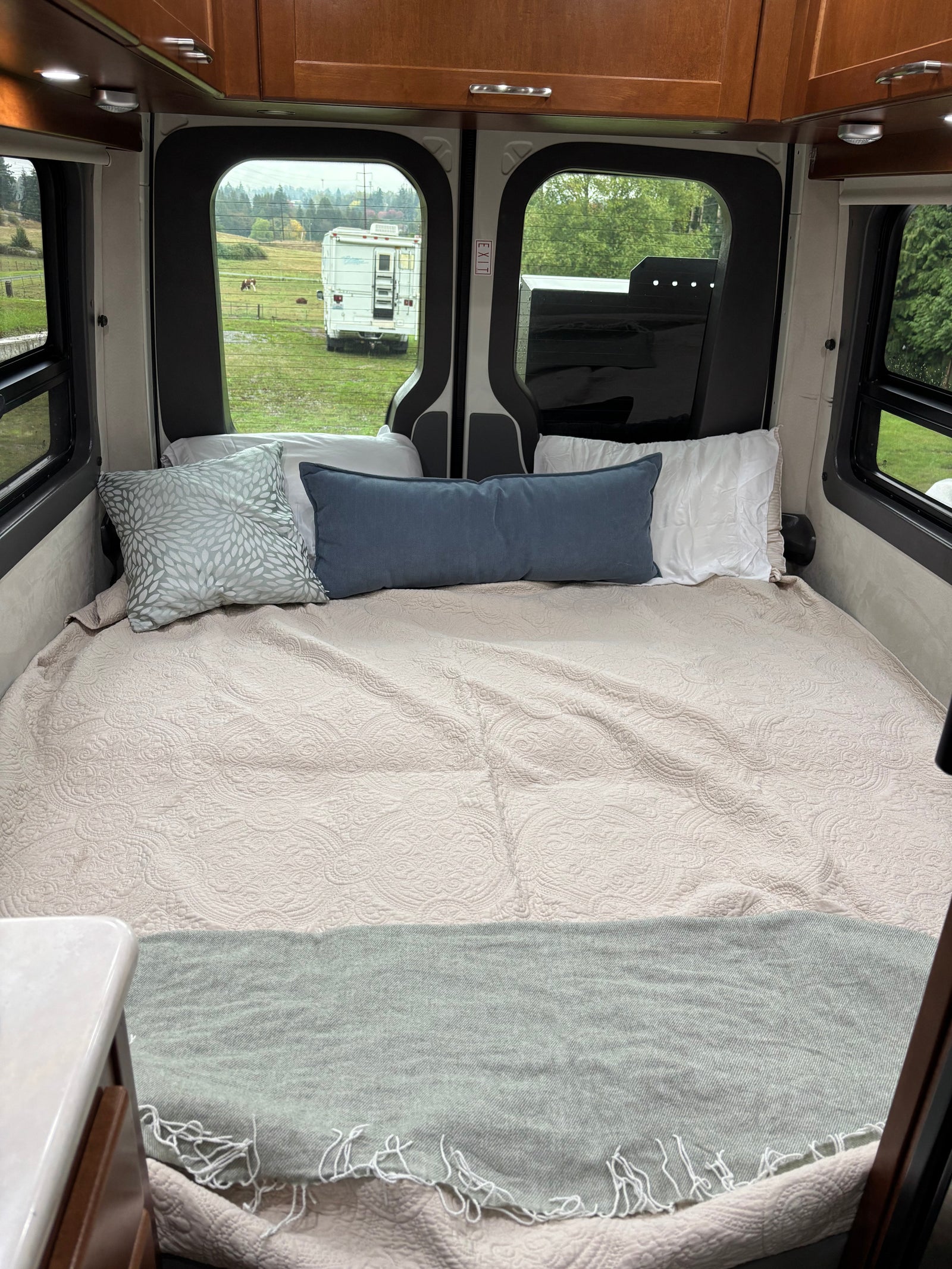 Cozy camper van bed for RV rental, featuring soft quilt, pillows, and serene views of a field and distant RV.