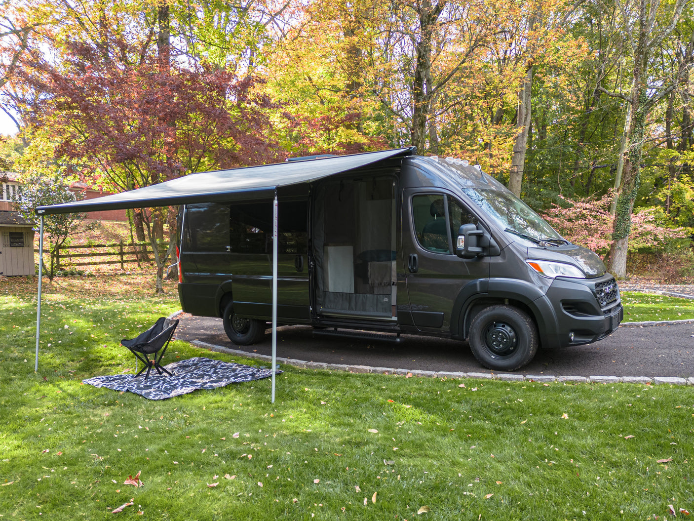 Dark gray camper van with awning extended, chair & blanket on grass in autumn. Ideal for your RV rental getaway.