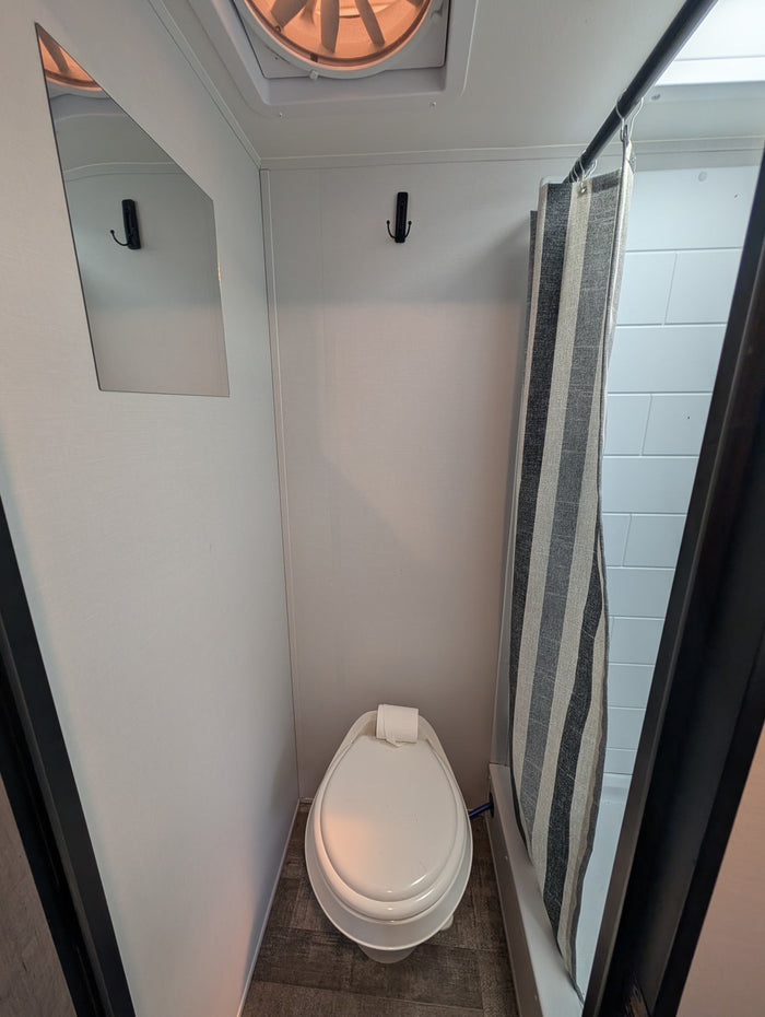 A compact RV rental wet bath with a toilet, striped shower curtain, wall mirror, and ceiling fan.