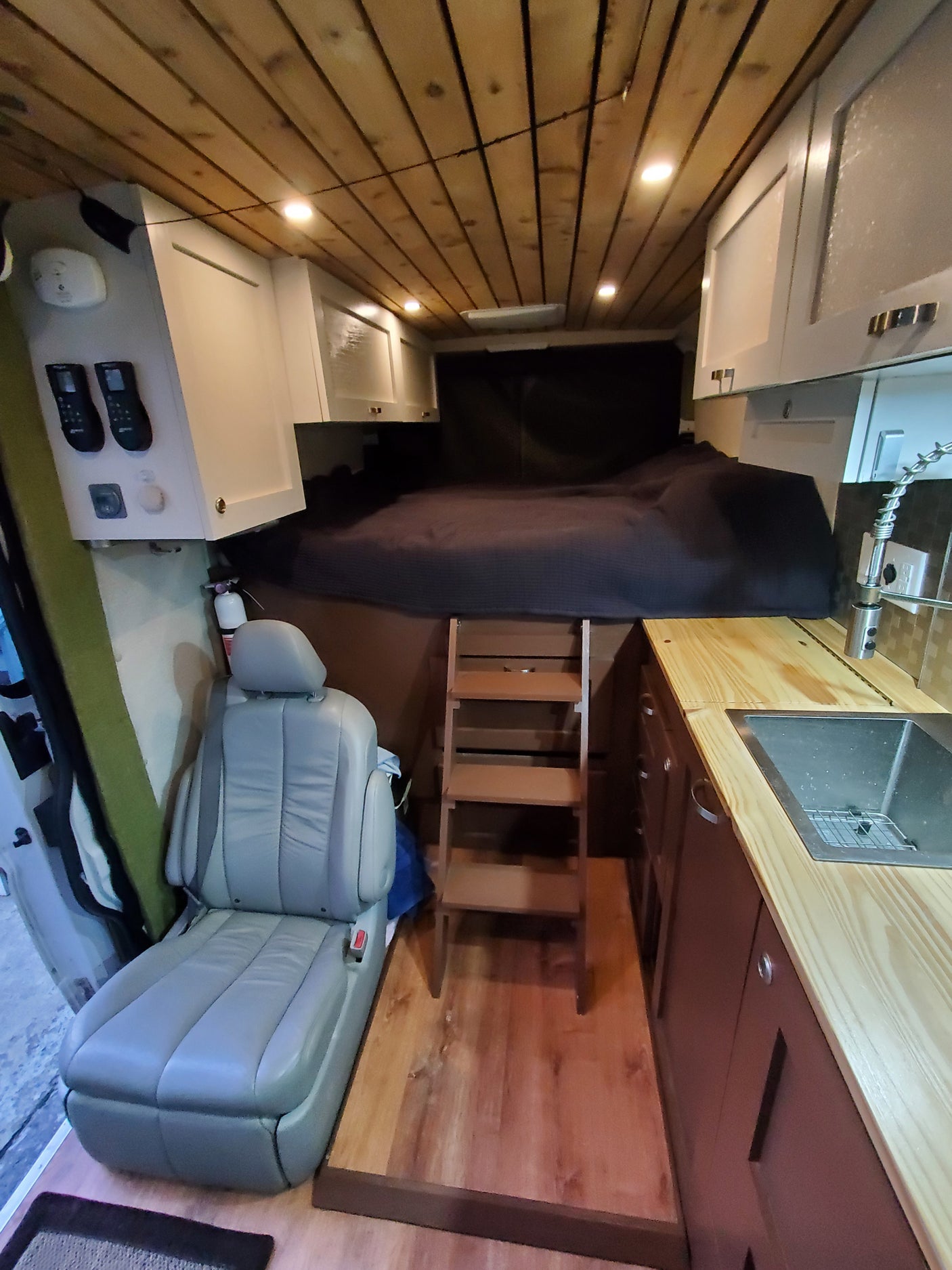 Inside a modern rv rental van: a cozy raised bed with ladder, compact kitchen with sink, light wood ceiling, and comfortable seating.
