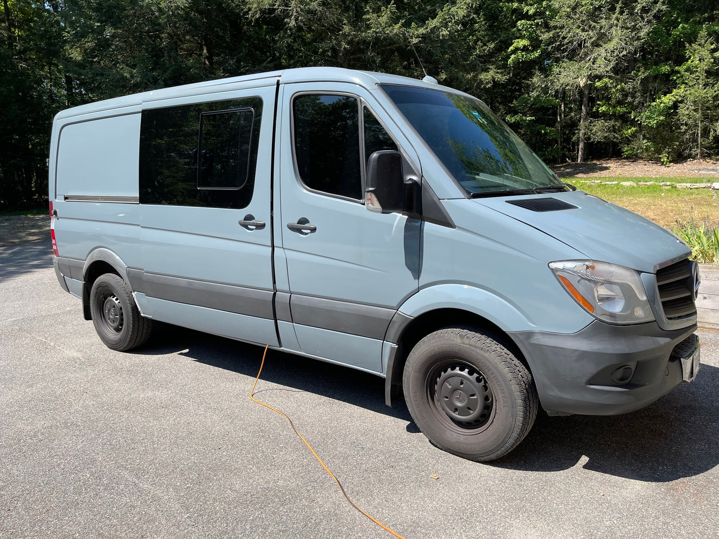 A light gray Mercedes-Benz Sprinter camper van, an ideal RV rental. Parked on asphalt outdoors with trees in the background.