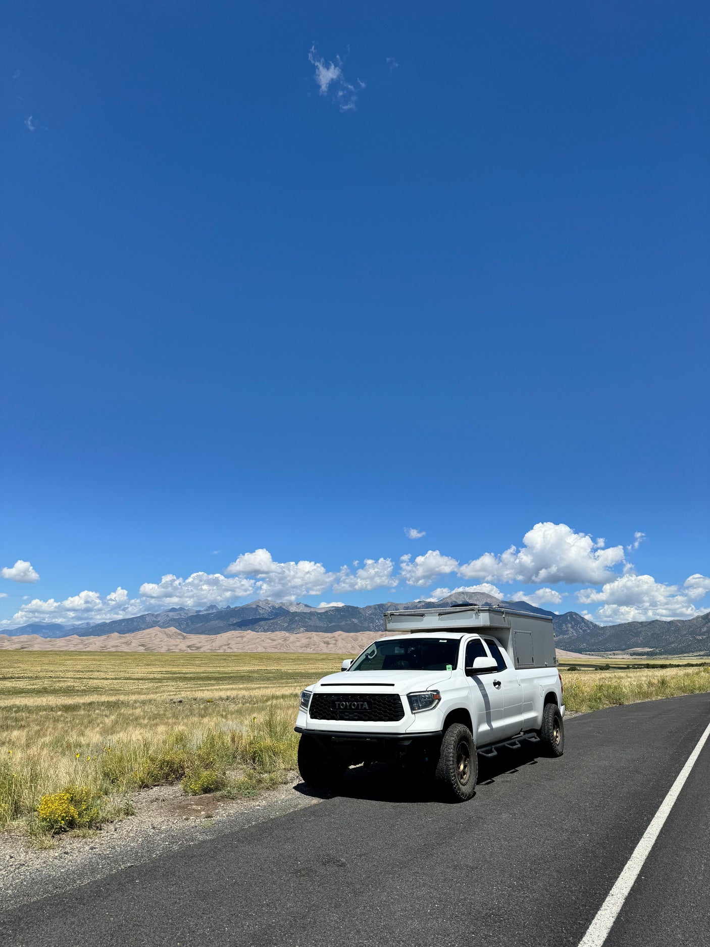 White Toyota Tundra pickup with an overland camper, reminiscent of an RV rental, parked on a scenic road with mountains and sand dunes.