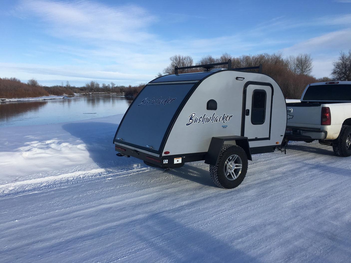 Bushwhacker teardrop RV rental parked on snow by a river, hitched to a white pickup truck.