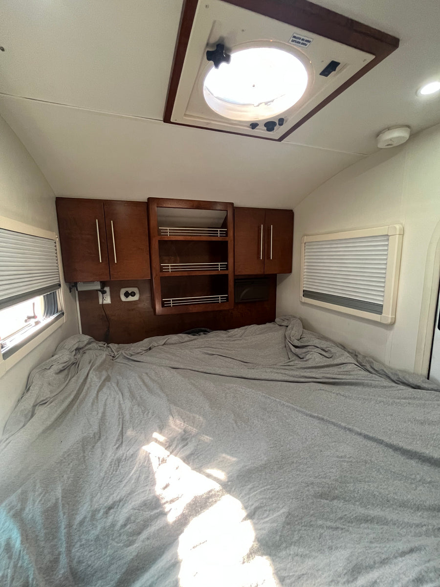 RV rental bedroom with a large bed, wooden overhead cabinets, windows with blinds, and a bright roof vent.
