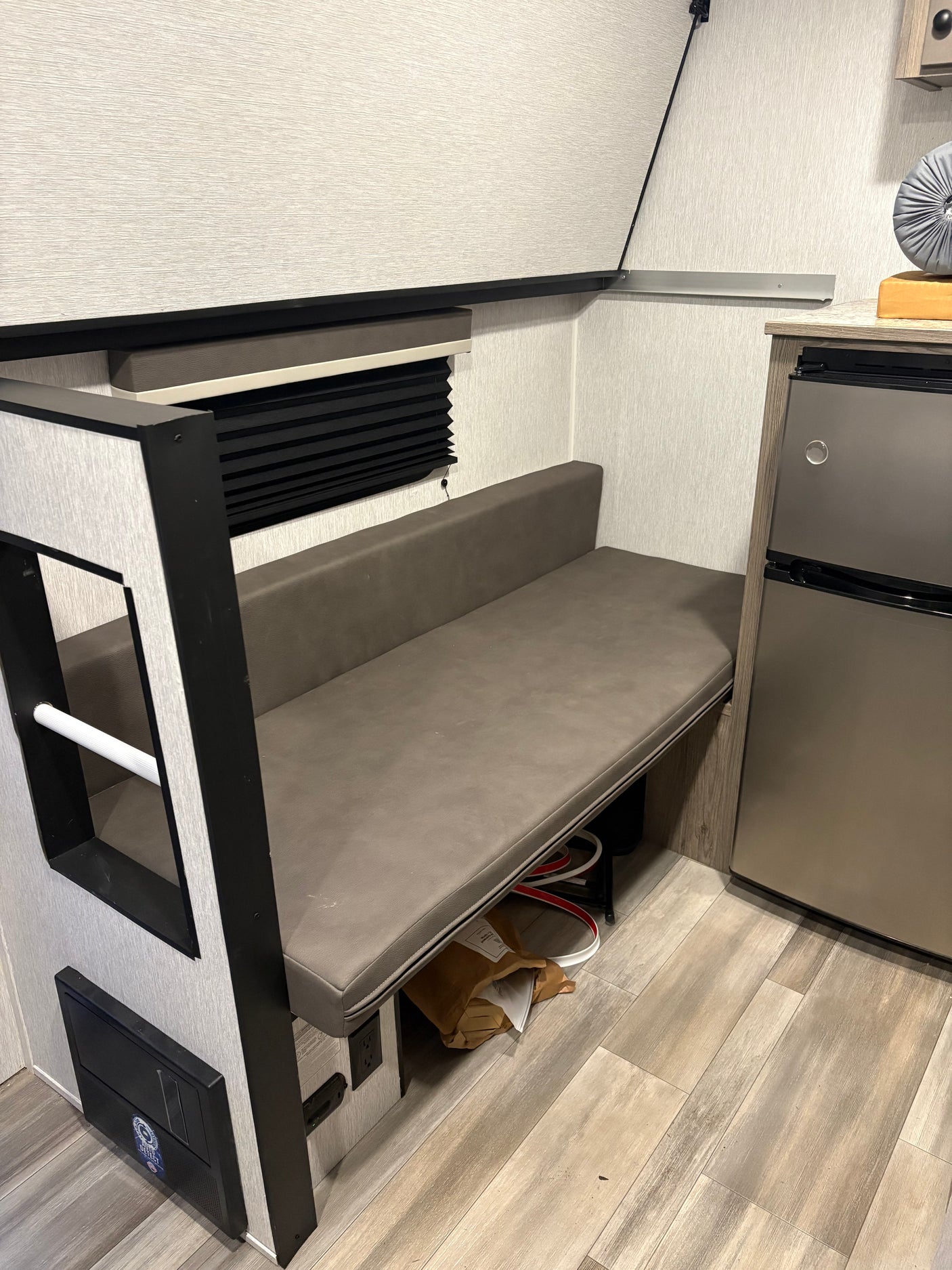 RV rental interior: cozy dinette with grey faux leather bench, window blind, and compact fridge. Light wood-look floor.