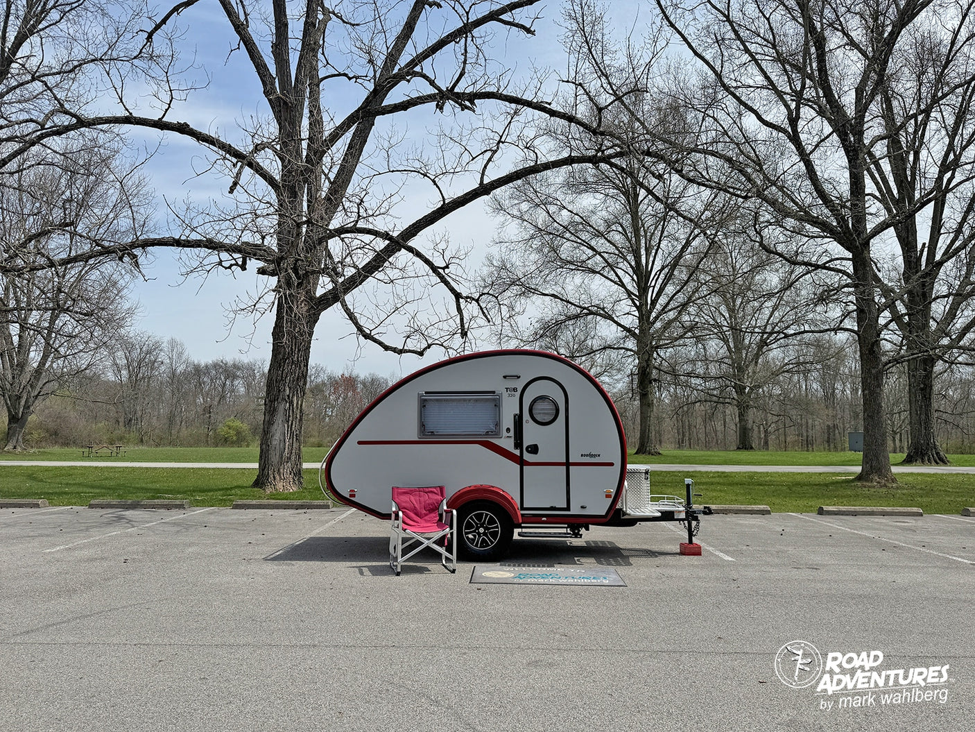 Enjoy your adventure! This small RV rental teardrop camper is perfectly set up in a park parking lot among bare trees.