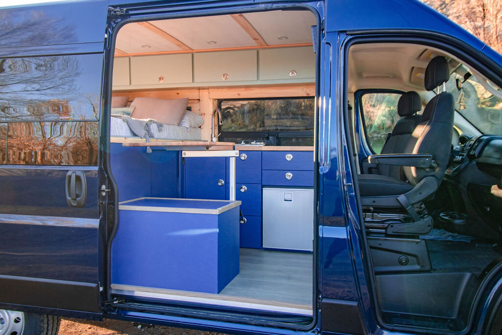 This blue camper van conversion shows a complete interior with bed, kitchen, storage, and front seats, ready for RV rental.