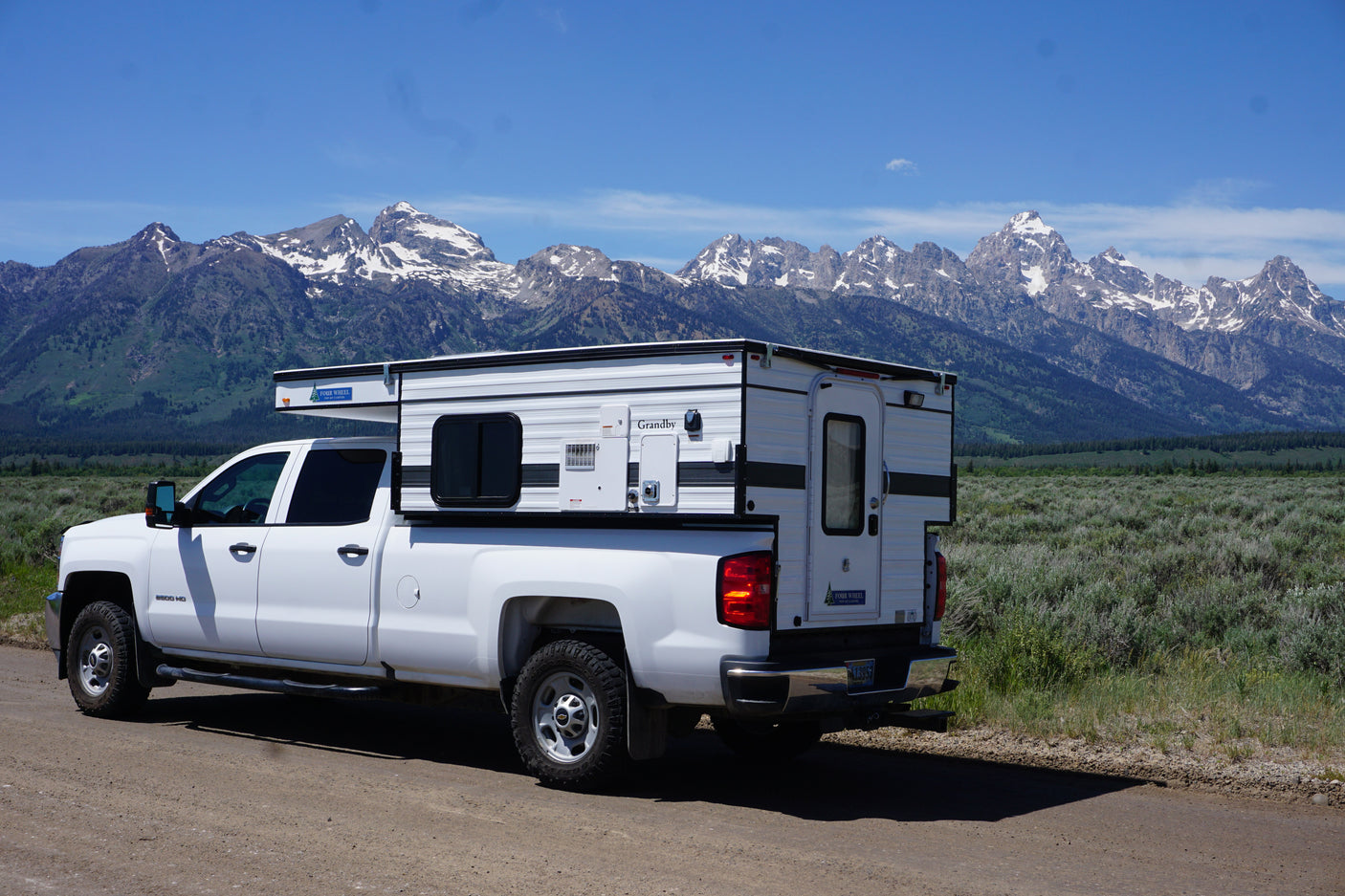 White pickup truck with a Four Wheel Pop-Up Camper RV rental, parked on a dirt road before grand, snow-capped mountains.