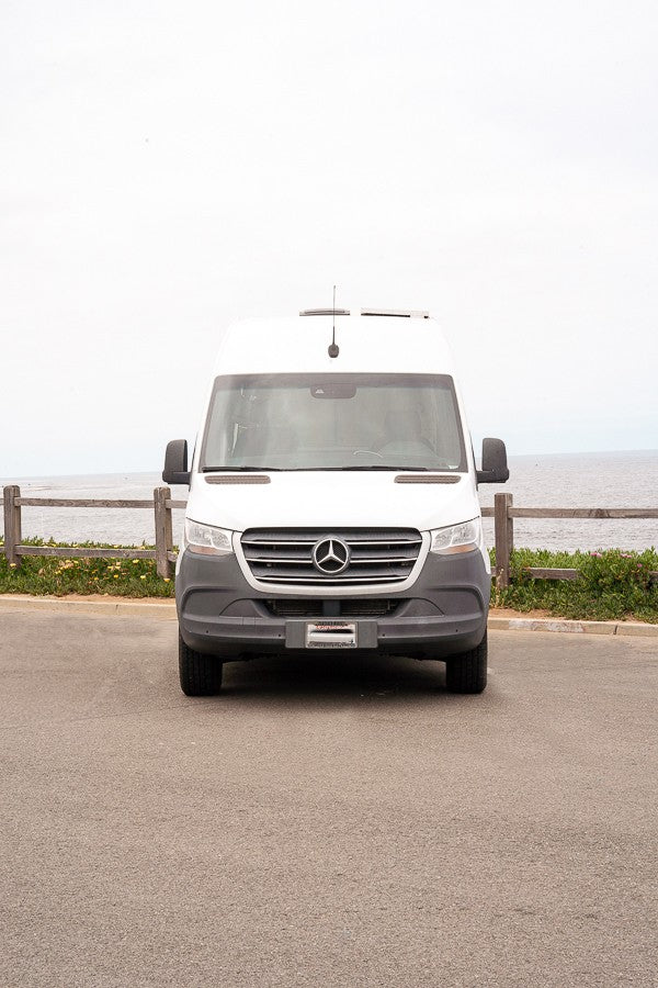 Front view of a white Mercedes-Benz Sprinter camper van, an RV rental, parked on asphalt by the ocean.